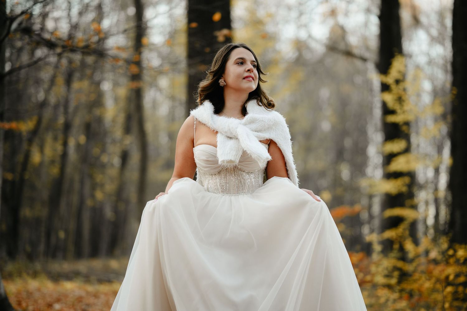 Bride in white wedding gown and fur shawl posing gracefully in an autumn forest setting.