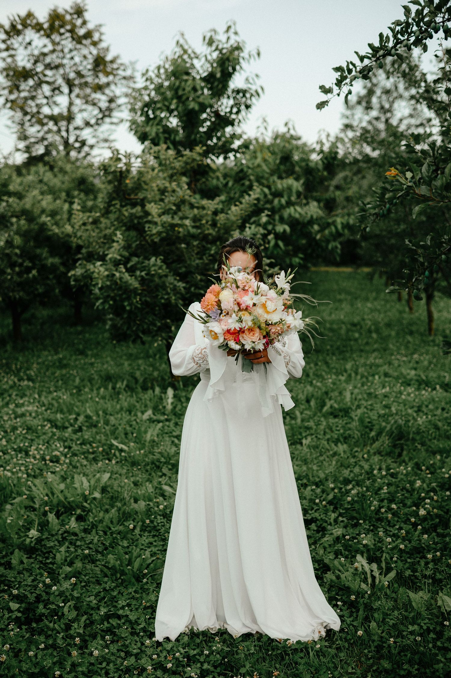 Bride in white long-sleeve gown holds colorful bouquet in lush green orchard.