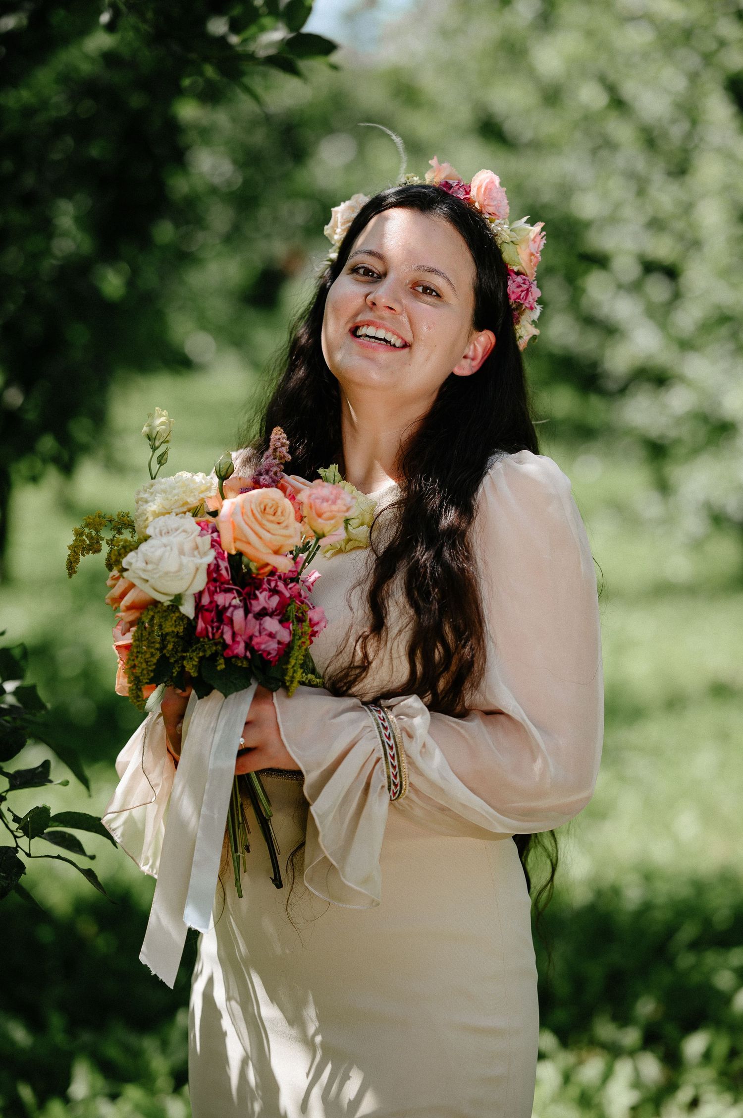 Smiling bride in white dress holding colorful bouquet with floral crown in sunny green garden.