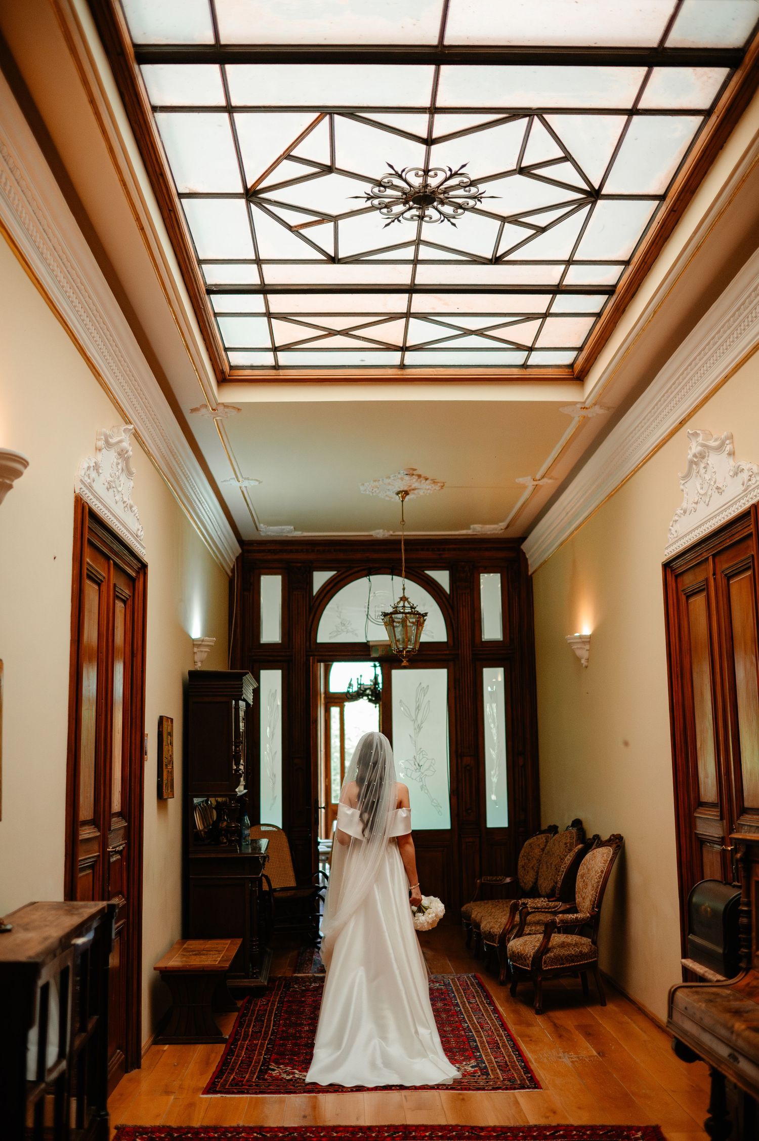 Bride in white gown with veil stands beneath ornate stained glass ceiling in grand foyer.