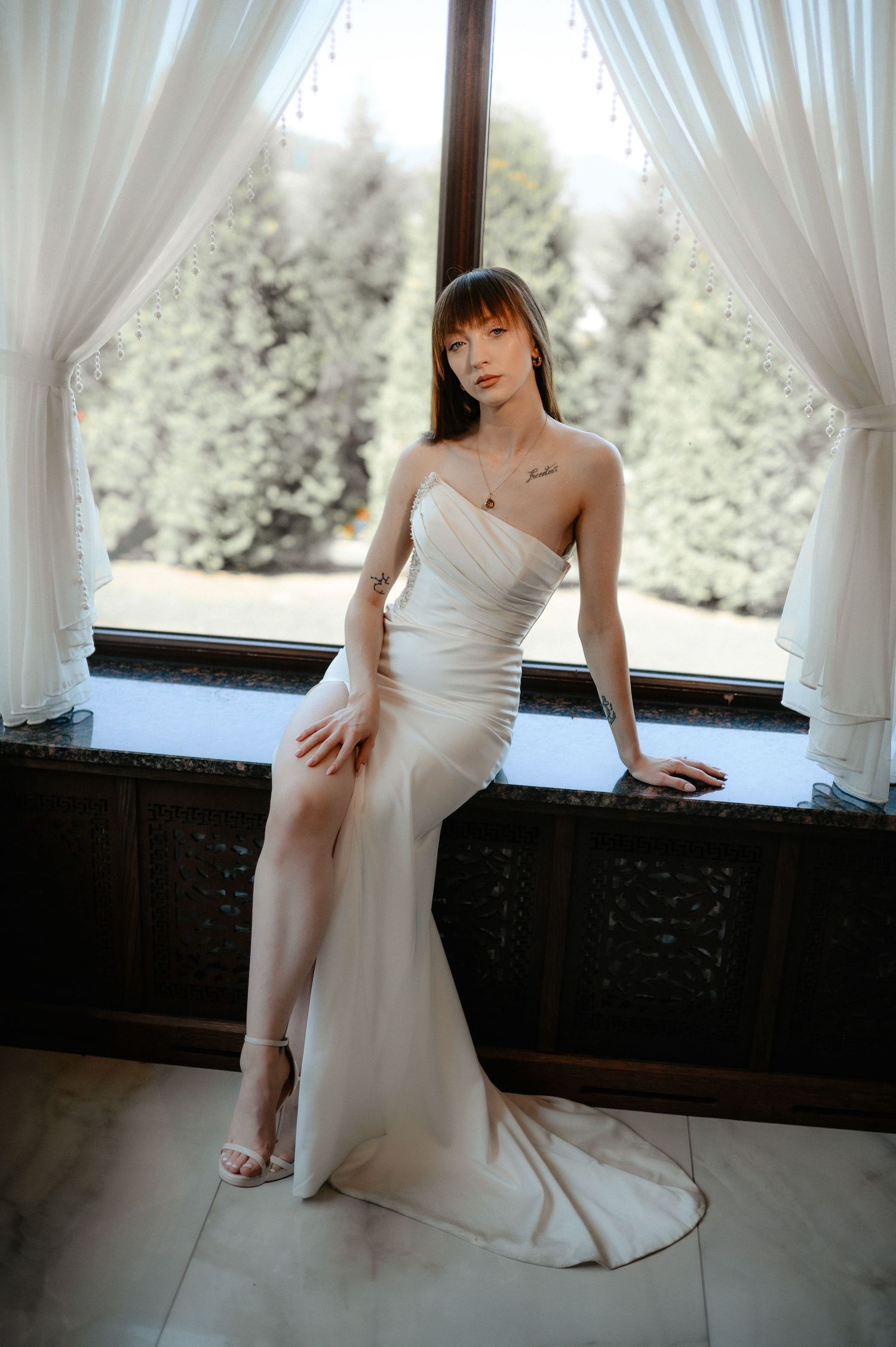 Bride in fitted strapless white gown with thigh slit poses gracefully by a sunlit window.