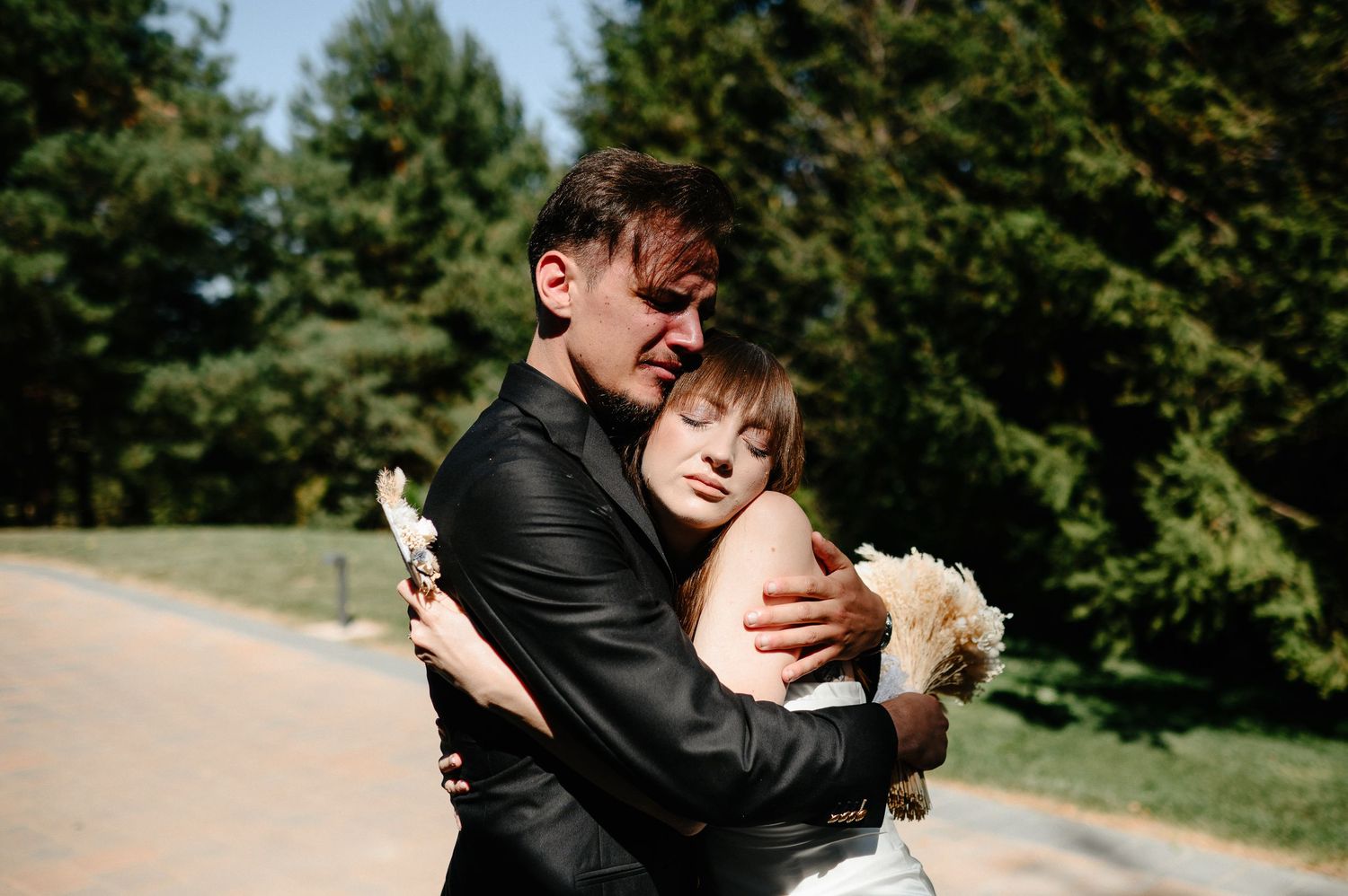 Emotional bride and groom embrace tightly outdoors surrounded by lush green trees.
