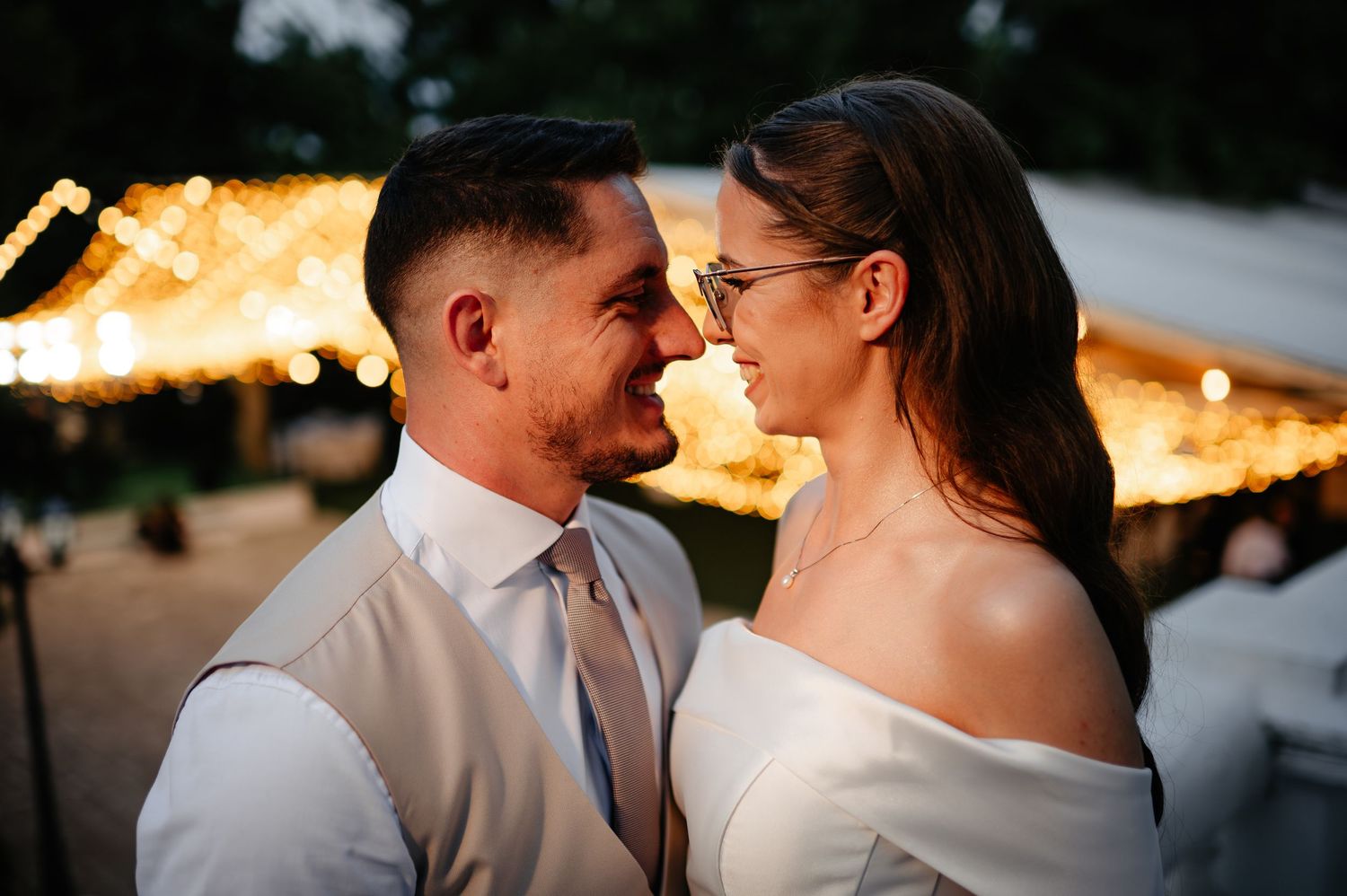 Newlyweds gazing lovingly at each other bathed in golden light from wedding reception string lights.