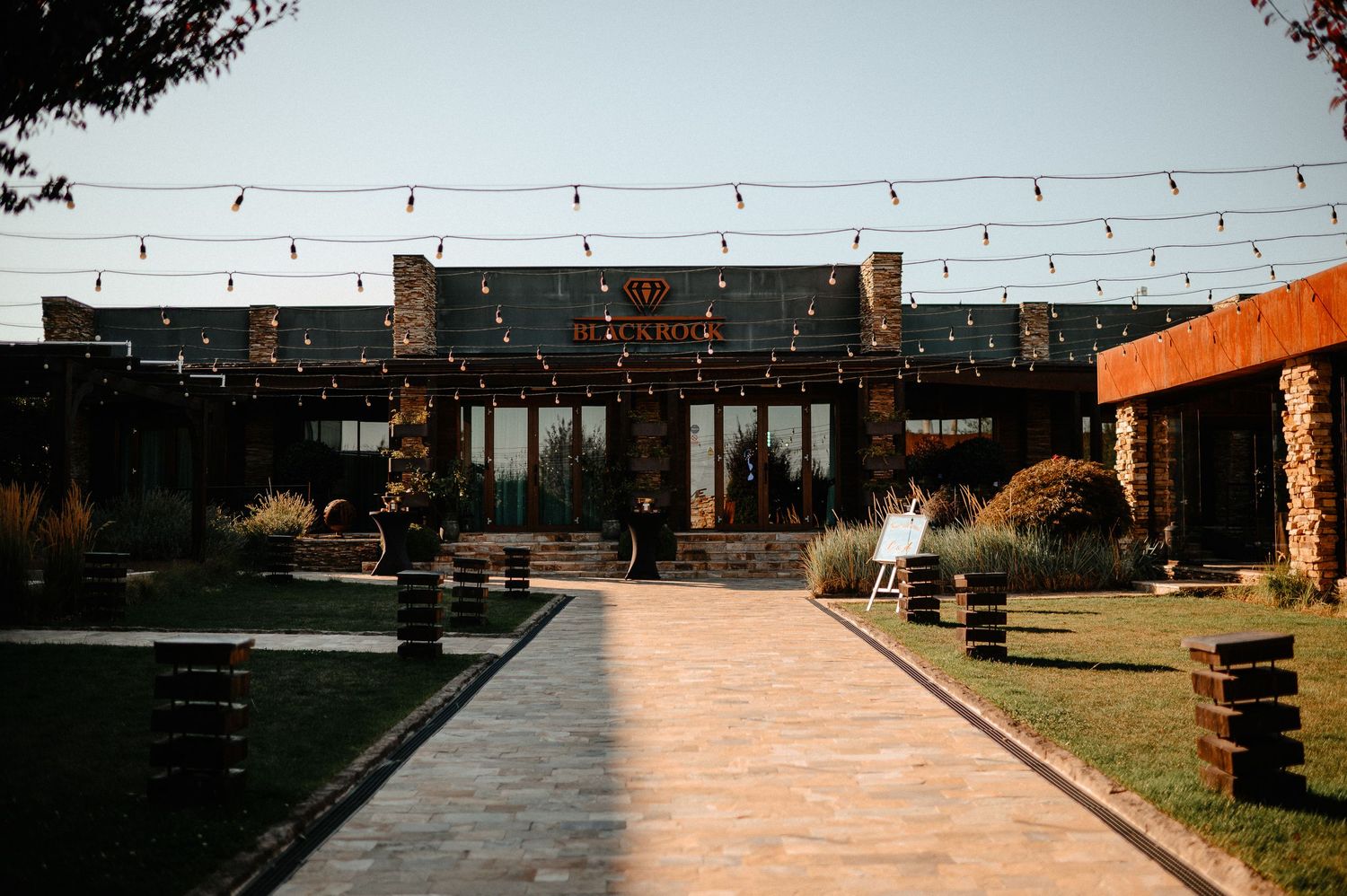 Rustic wedding venue exterior at golden hour with string lights and brick pathway.