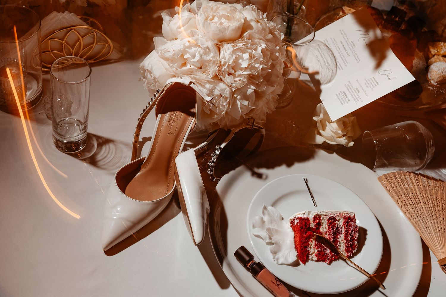 Elegant wedding table setting featuring white bridal heels, peony bouquet, red velvet cake slice, and menu card.