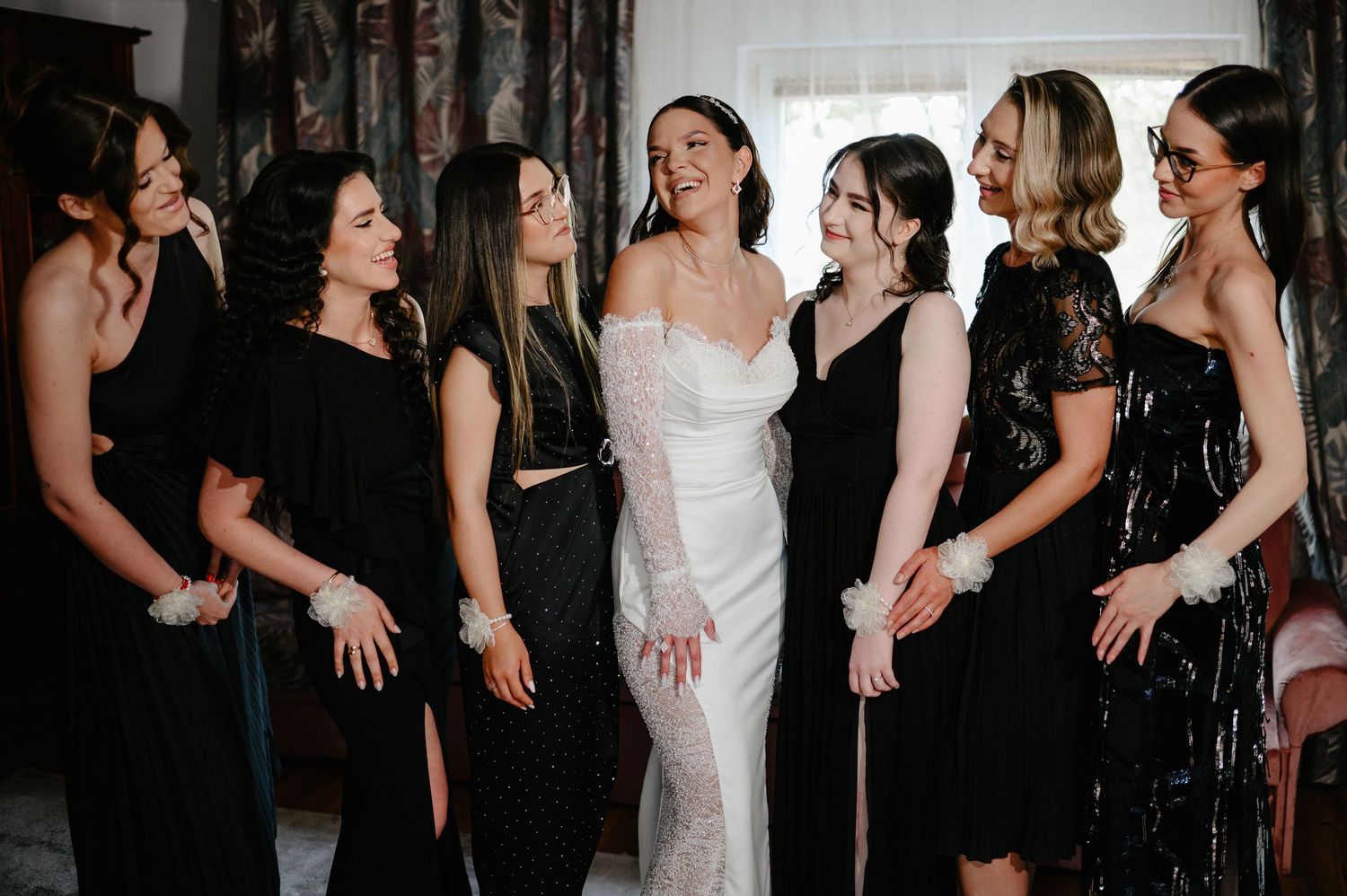 Bride in white dress laughs with six bridesmaids wearing coordinated black dresses indoors.