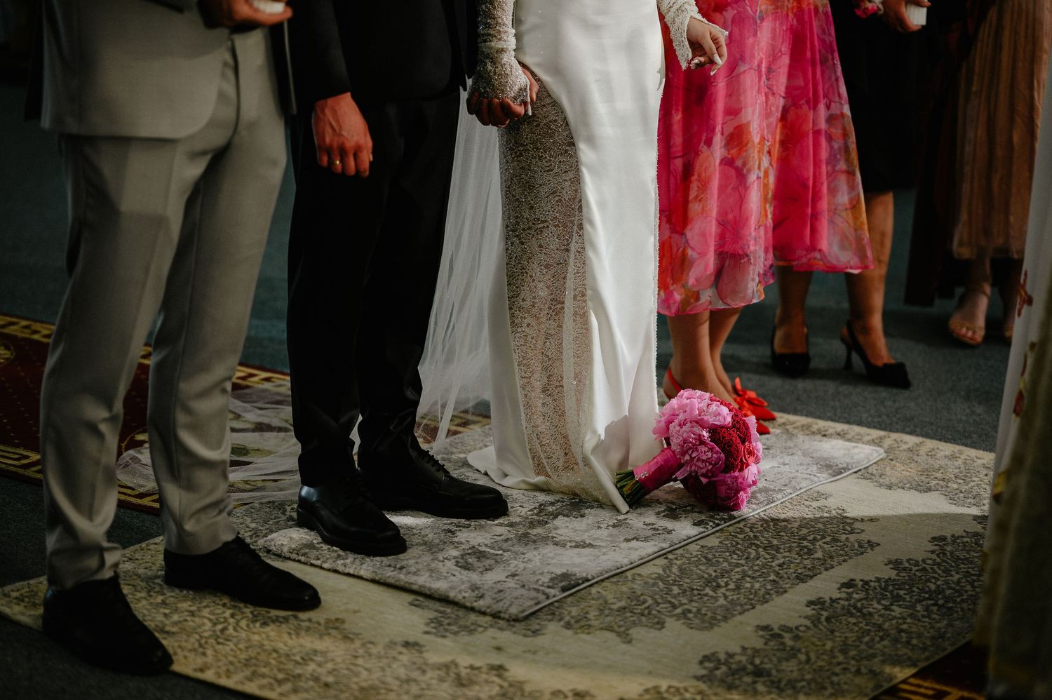 Bride and groom stand on decorative rug during wedding ceremony with pink bouquet on floor.