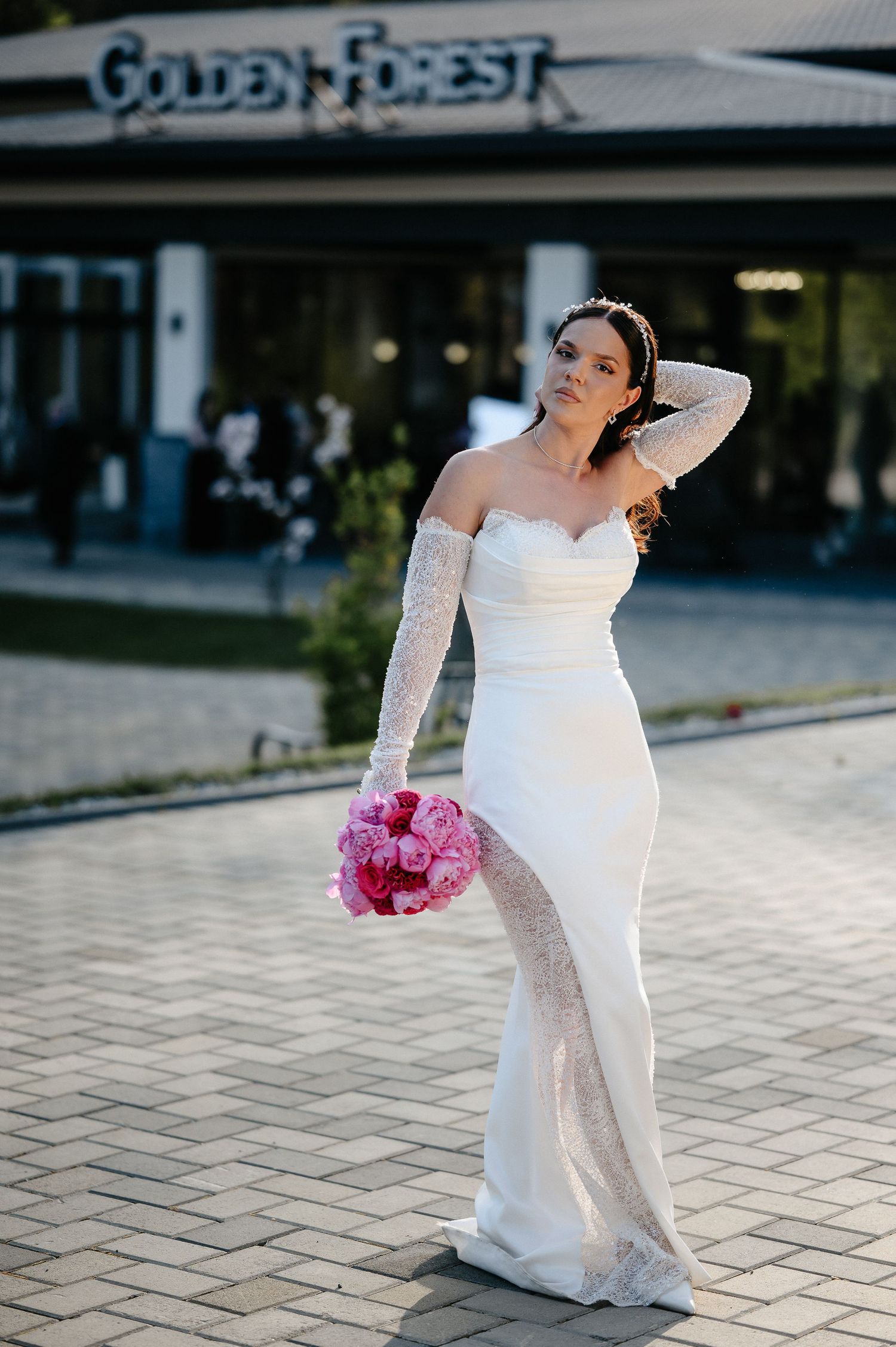 Beautiful bride posing in fitted white wedding gown with lace sleeves outside Golden Forest, holding pink peonies.