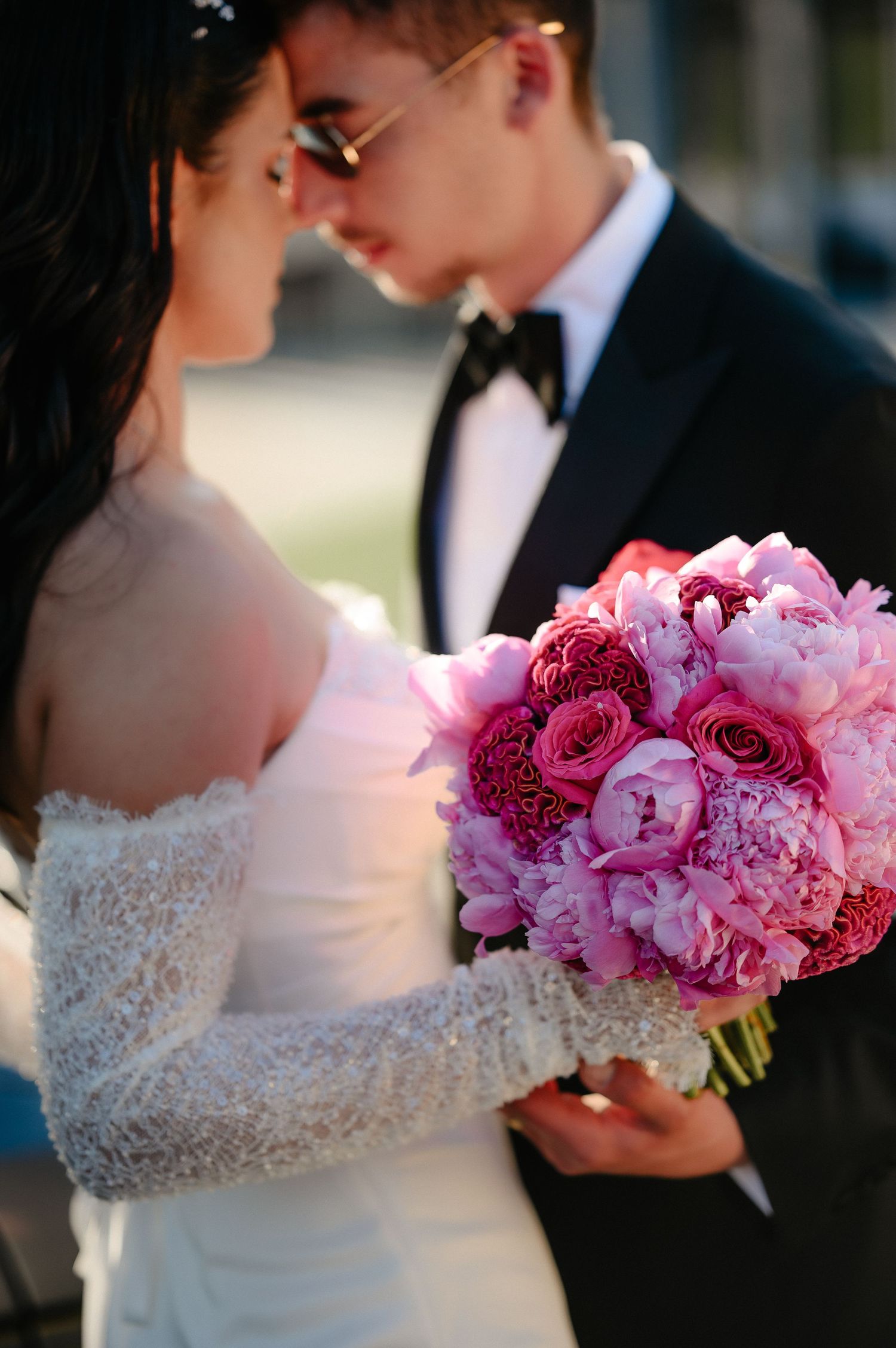 Close-up of bride and groom touching foreheads, bride holding lush pink peony and rose bouquet.