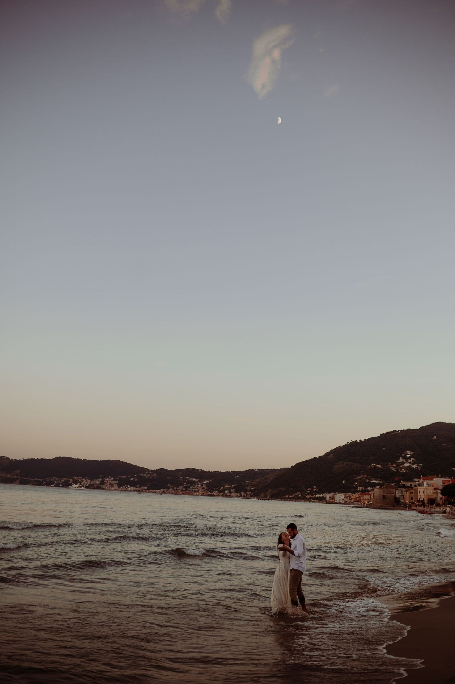 Couple embracing at the shoreline during sunset with a crescent moon above.