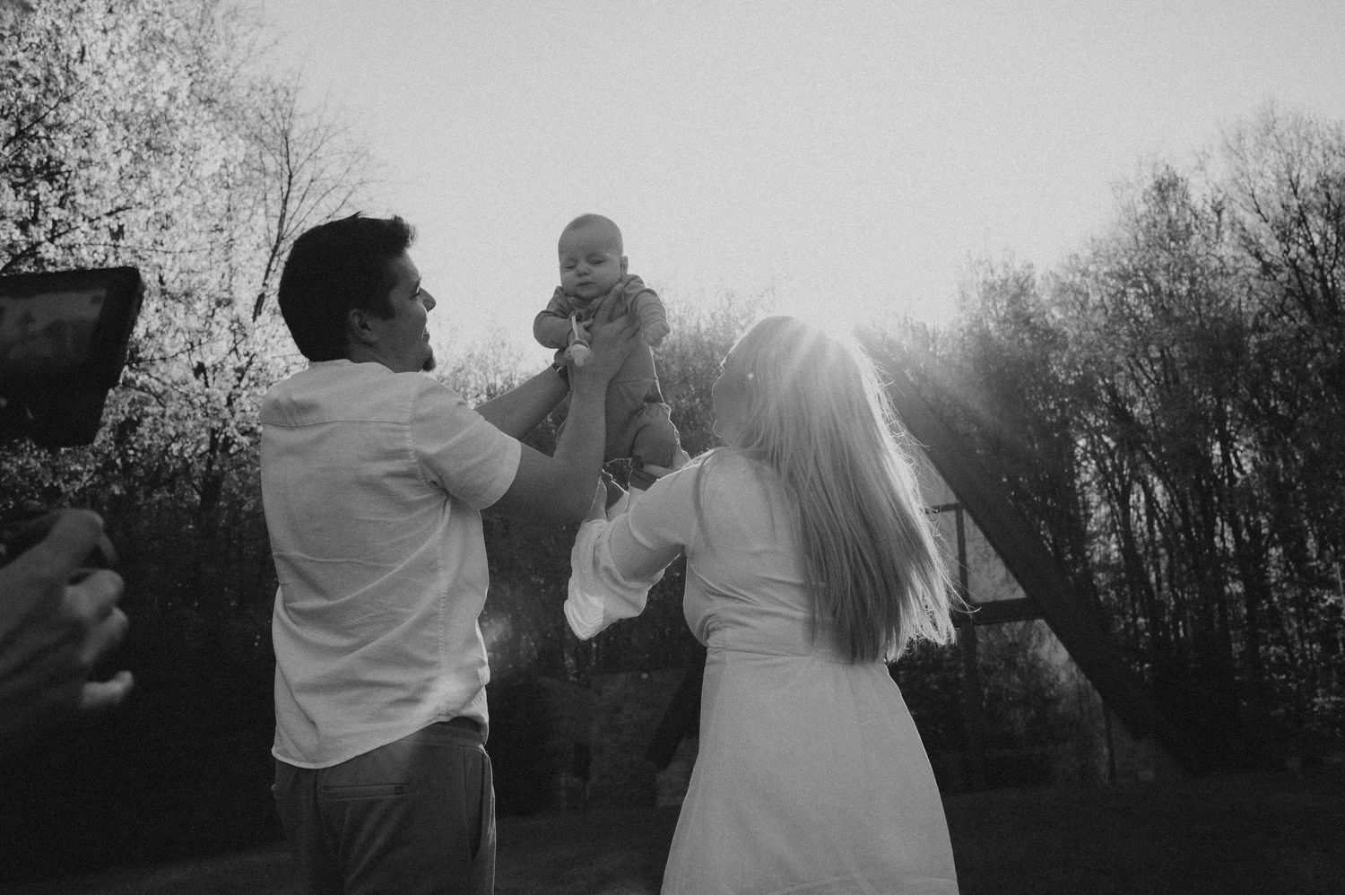 Black and white photo of couple lifting their baby together outdoors with sunlight streaming behind them.