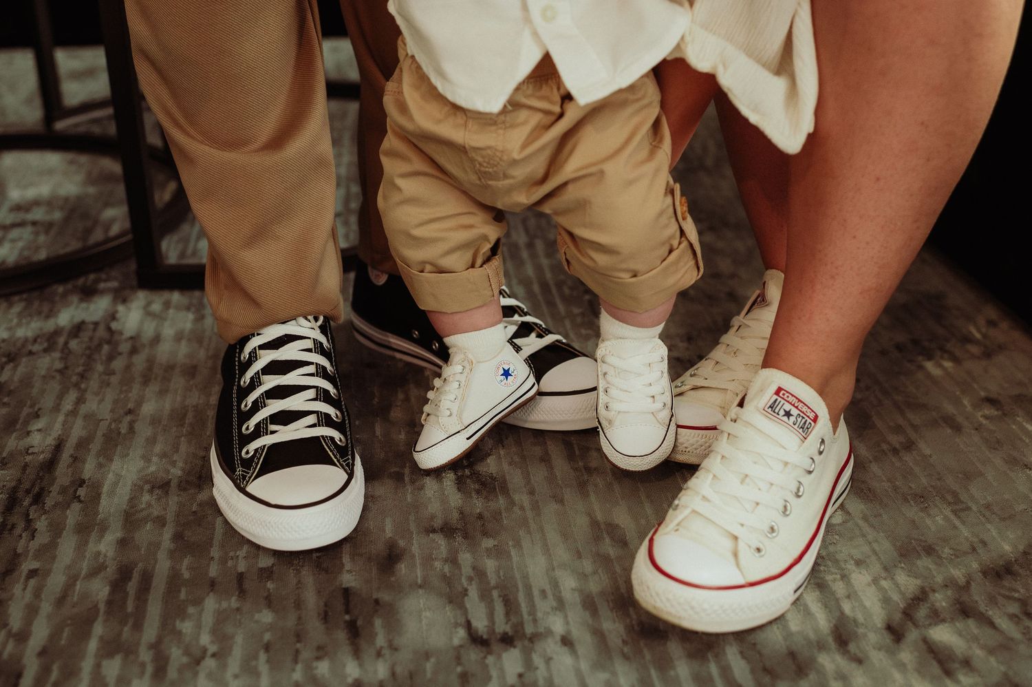 Three generations wear matching Converse sneakers, showcasing black, white, and classic styles on a wooden floor.