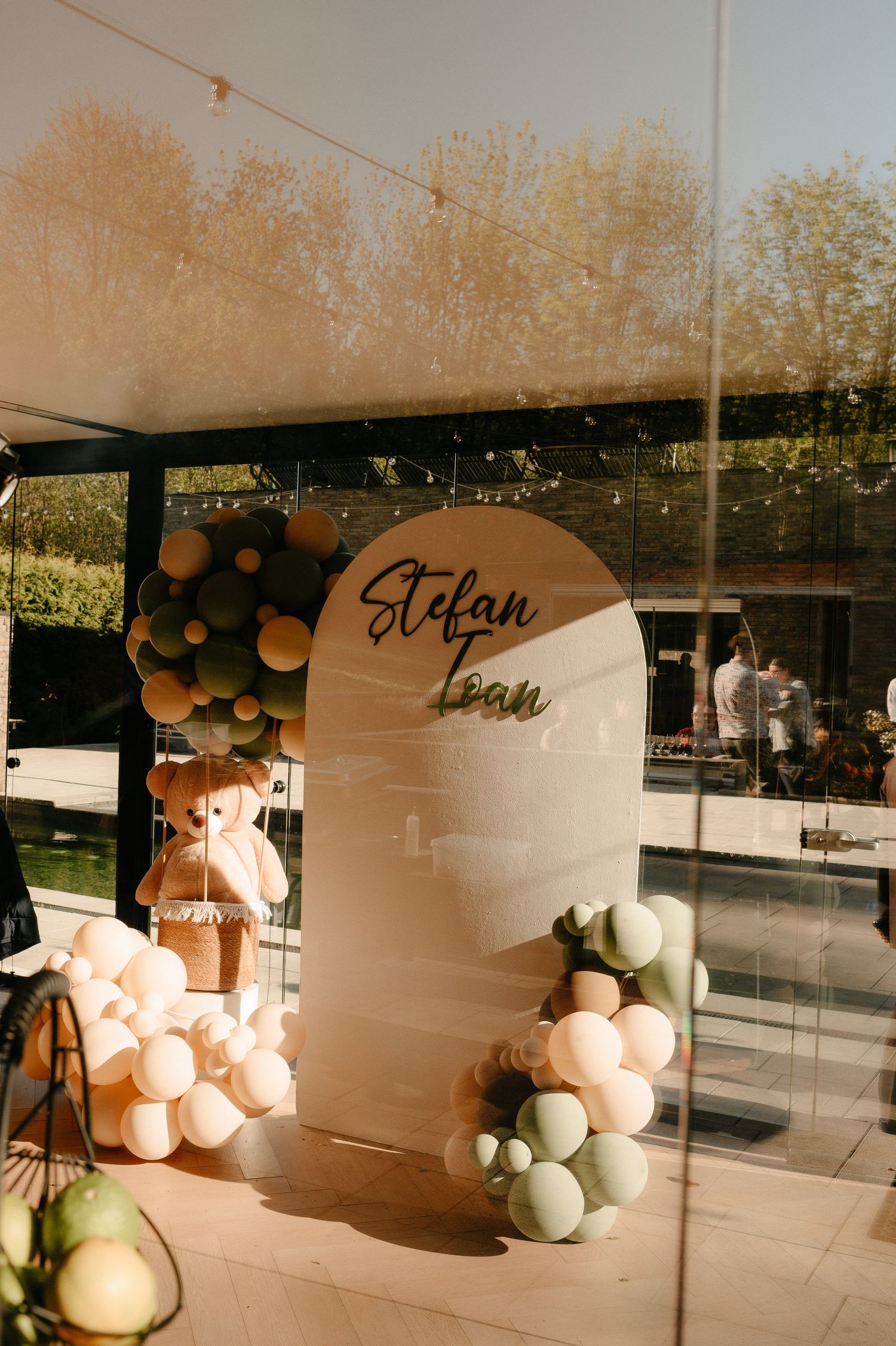 Elegant baby christening backdrop with Stefan Ioan name sign, teddy bear, and sage green balloon decor.