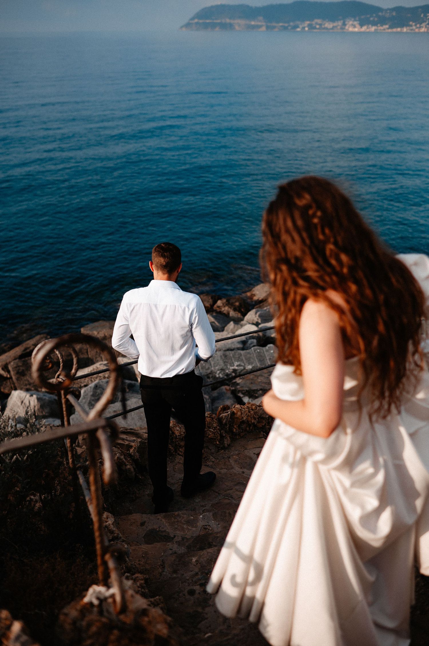 Bride in white gown watching groom gaze at deep blue Mediterranean sea from rocky steps.