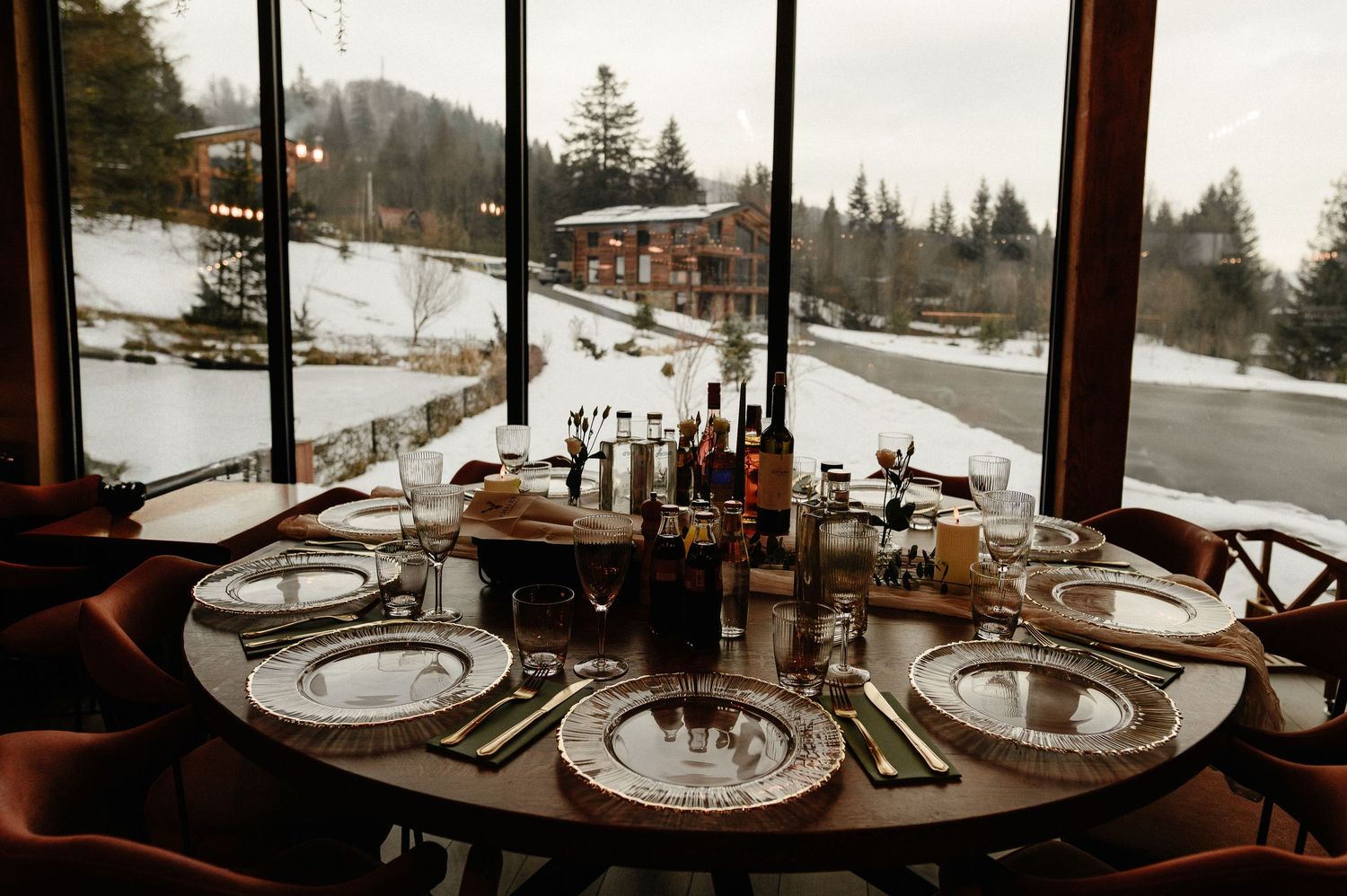 Elegant round dining table set with silver charger plates overlooking a snowy winter landscape through floor-to-ceiling windows.