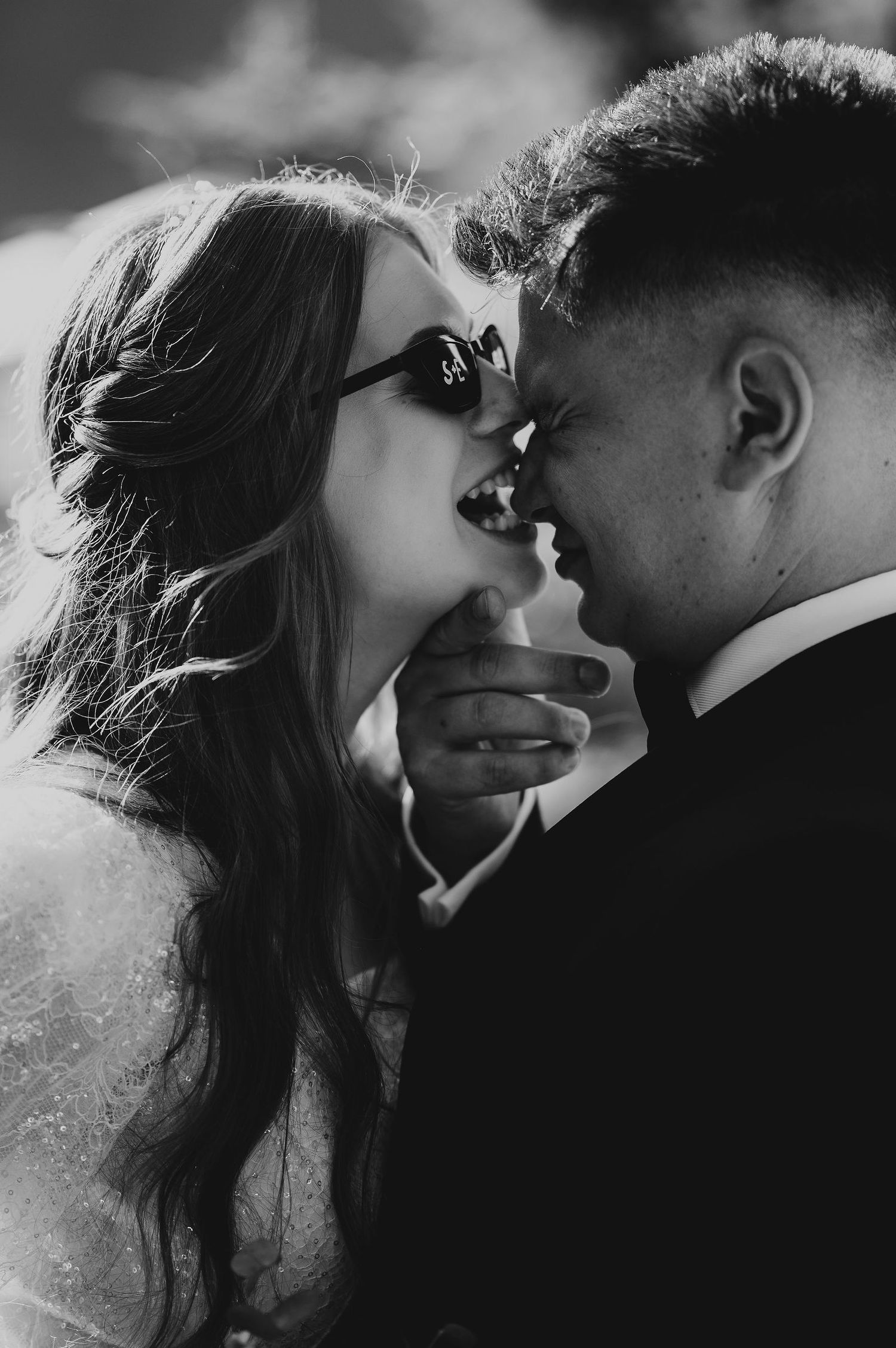 Black and white photo of smiling bride with sunglasses and groom sharing intimate moment.
