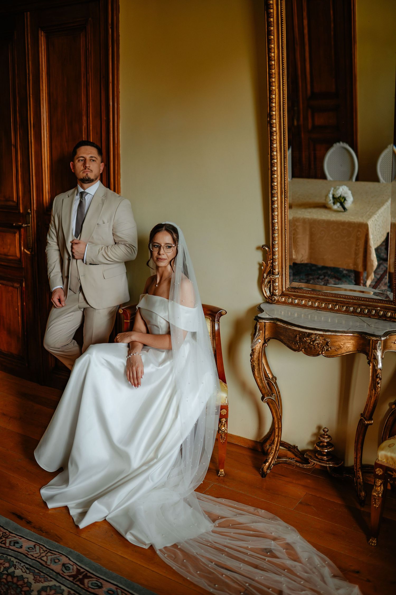 Bride in white gown with veil seated beside groom in beige suit in ornate room.