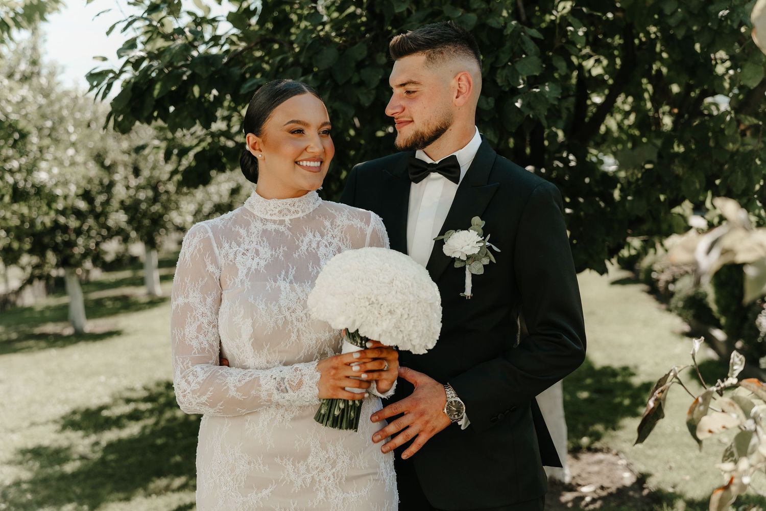 Happy bride and groom posing together outdoors, bride holding white bouquet in lace dress.