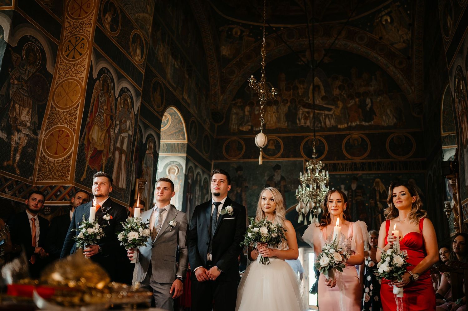 Wedding party at Orthodox church altar with ornate frescoed walls and chandelier.