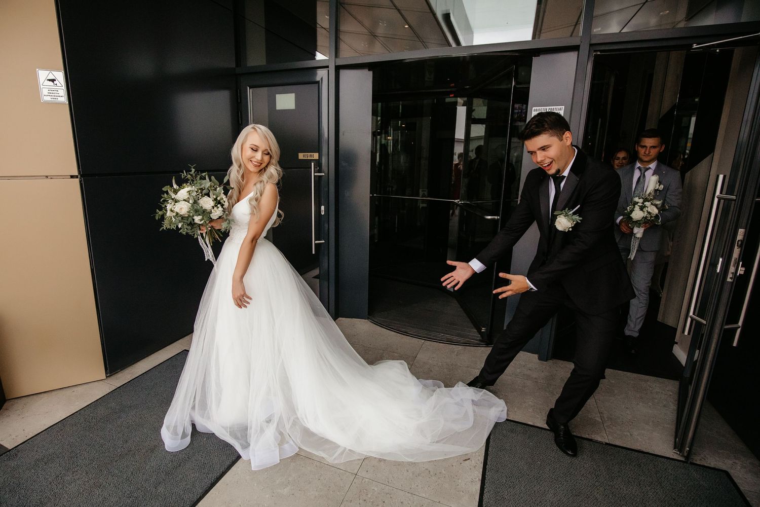 Bride in white tulle gown with bouquet and groom in black suit exiting building through glass doors.