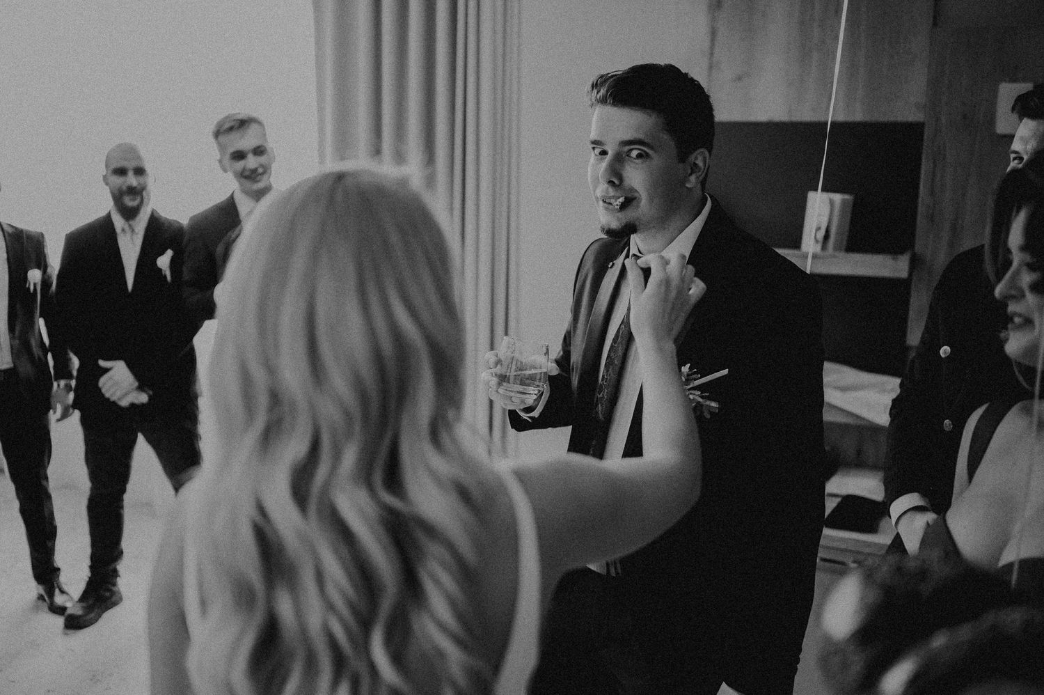 Groom adjusting his tie while groomsmen laugh and chat in a stylish black and white wedding preparation photo.