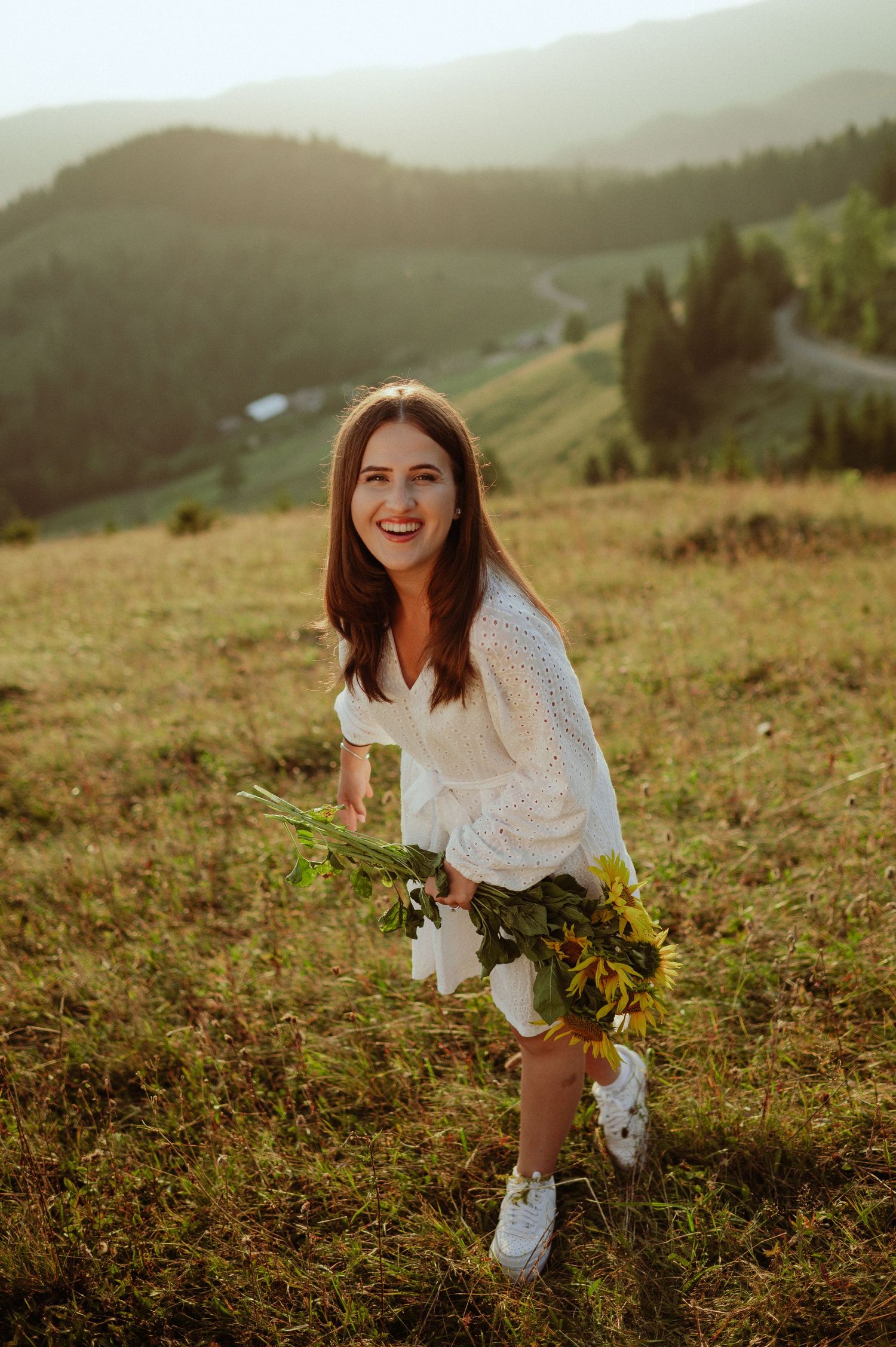 Smiling brunette woman in white dress holding sunflowers in a mountain meadow at sunset.