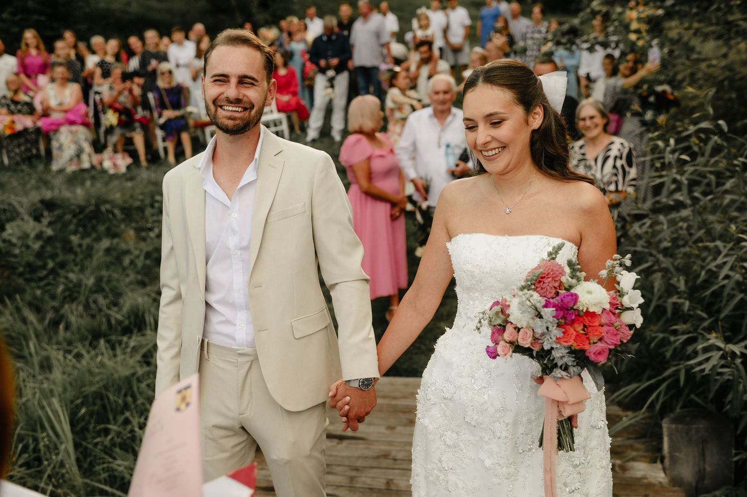 Smiling newlyweds walk hand in hand past guests after outdoor wedding ceremony with colorful bouquet.