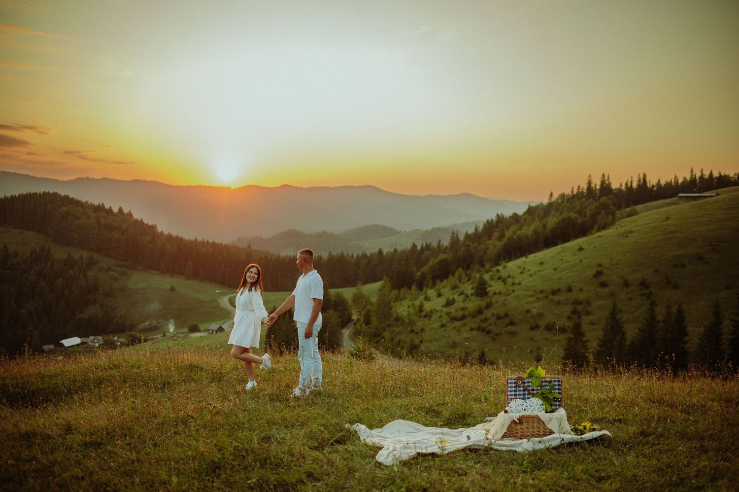 Couple in white outfits walking hand-in-hand at golden sunset on a green mountain hillside.