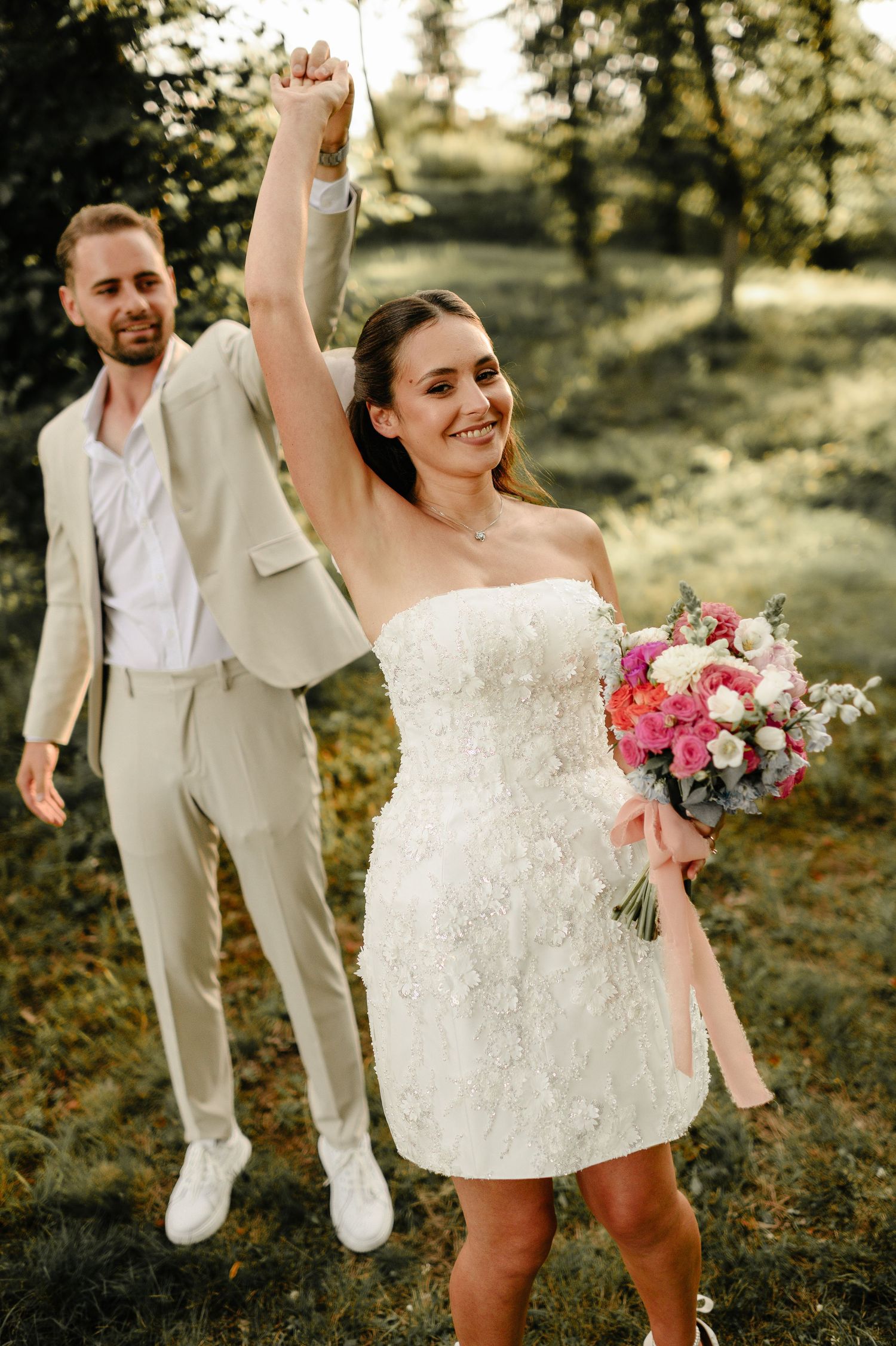 Joyful bride in short white lace dress raises hand in celebration with groom in beige suit outdoors.