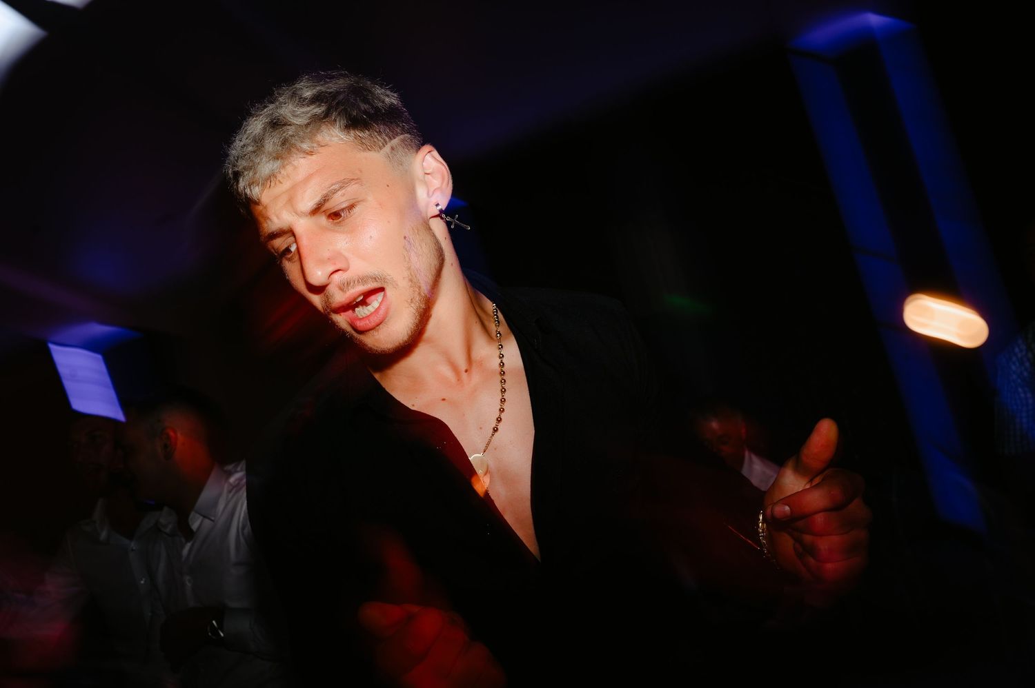 Young man with bleached hair dancing at nightclub event under dramatic blue and white lighting.