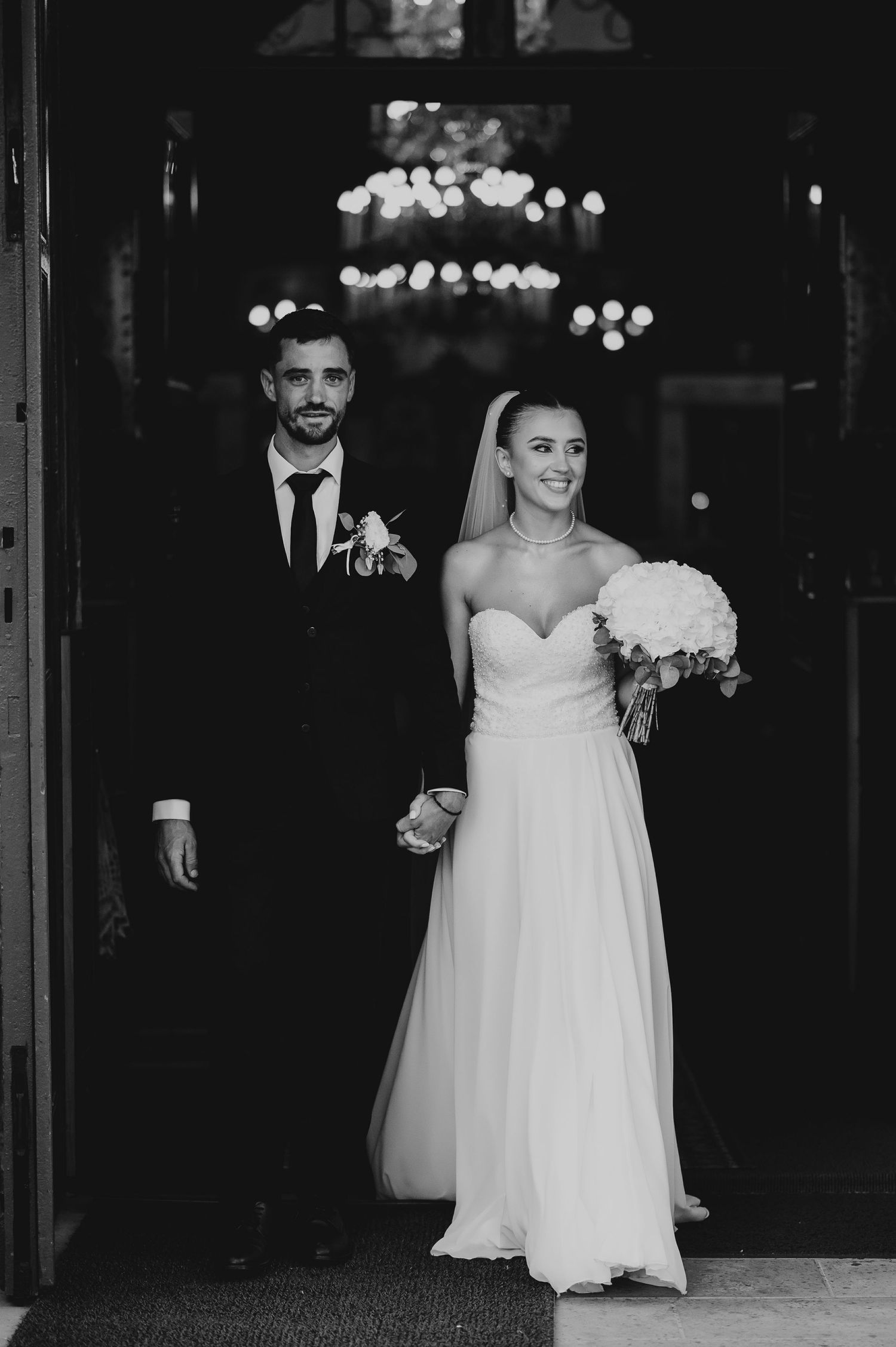 Black and white photo of newlyweds exiting church, bride in strapless gown holding bouquet, groom in suit.