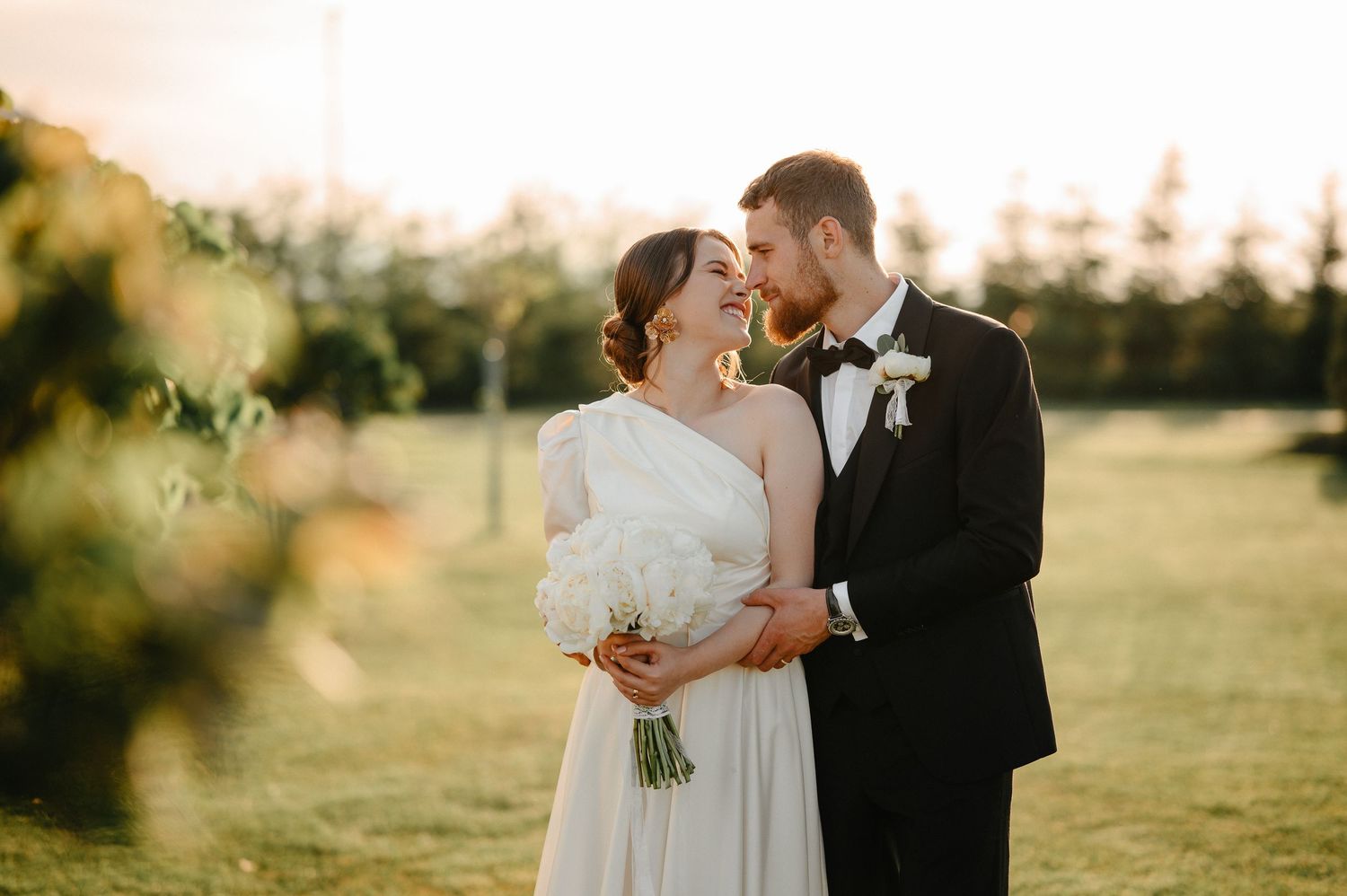 Bride and groom share a romantic golden hour moment outdoors, bride holding white bouquet in elegant wedding attire.