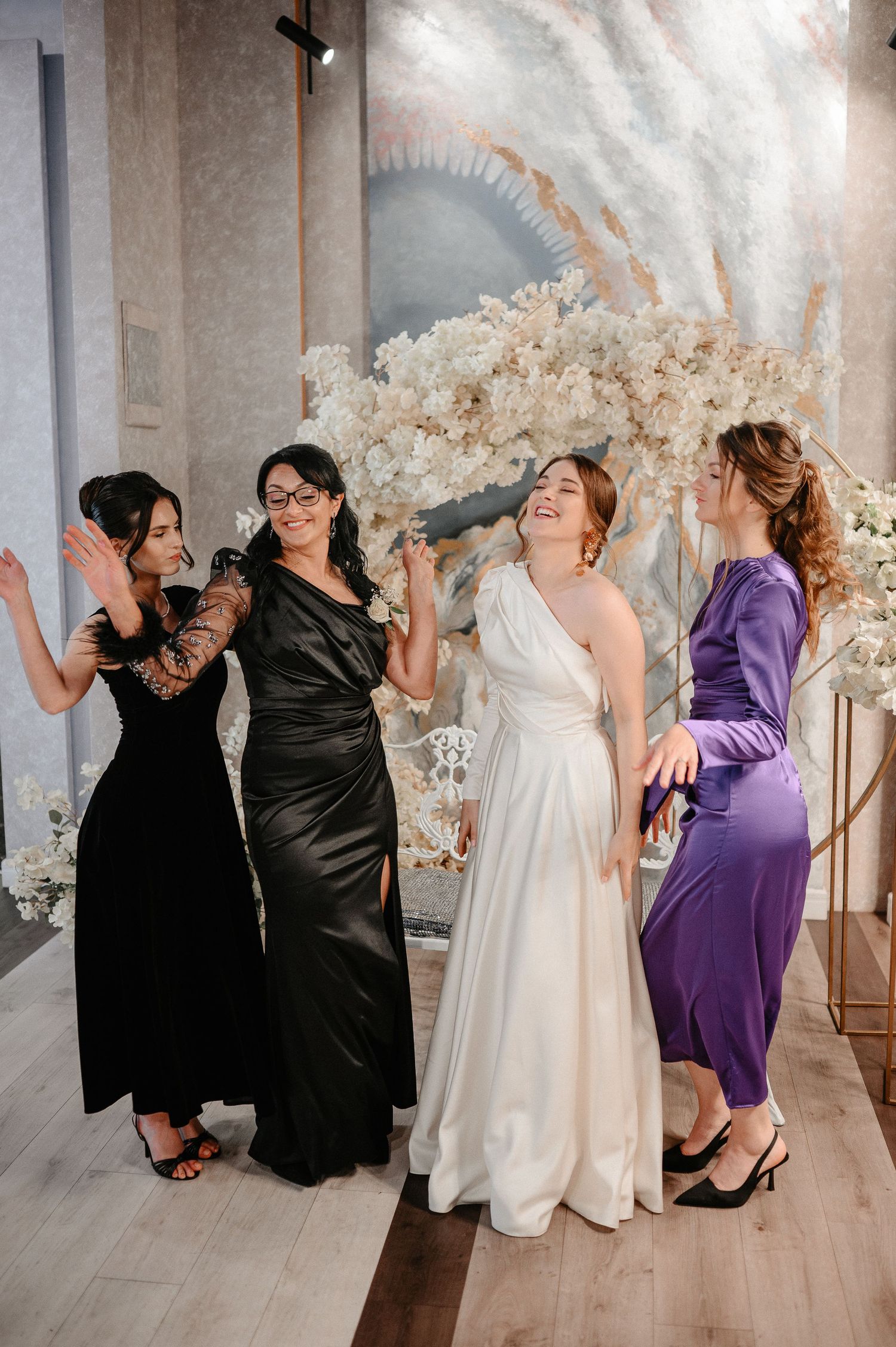 Bride in white gown laughs joyfully with three bridesmaids in black and purple dresses near floral arch.