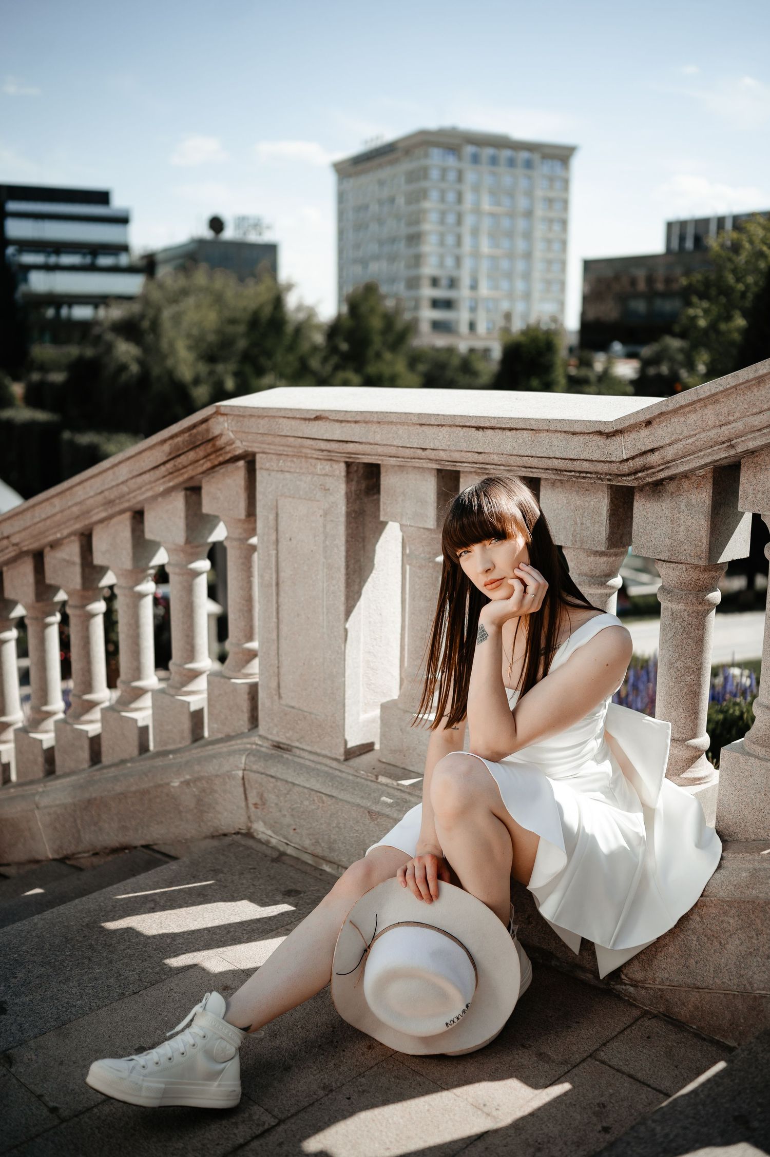 Young woman in white dress and sneakers sits on stone balustrade steps holding a wide-brim hat in an urban setting.