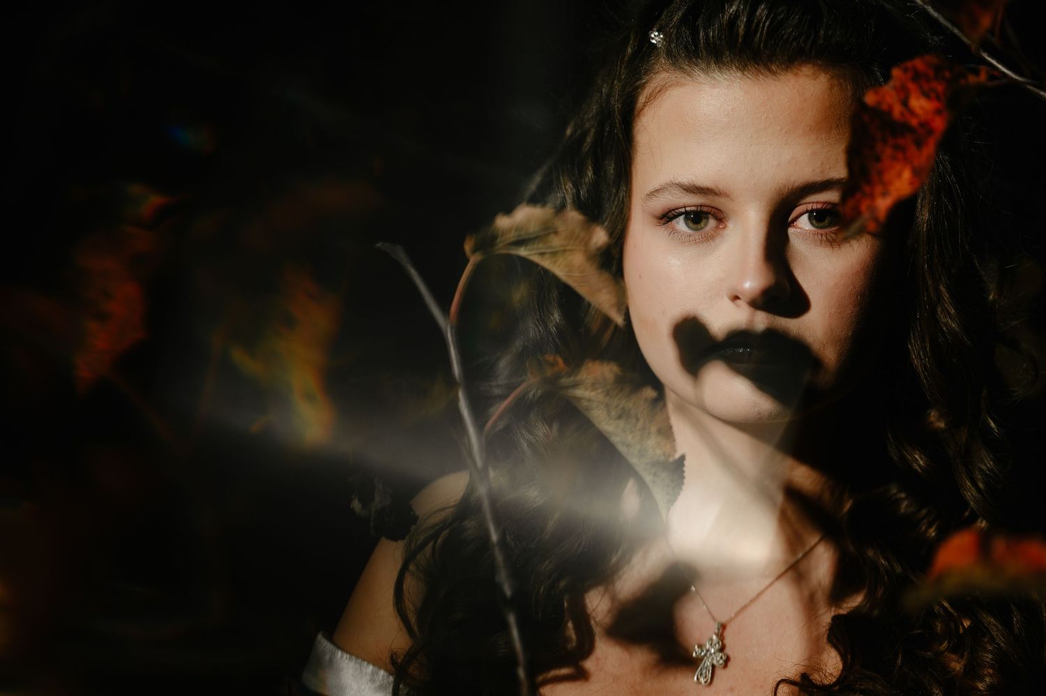 Mysterious young woman in Halloween costume with dark lipstick and dramatic lighting through branches at night.
