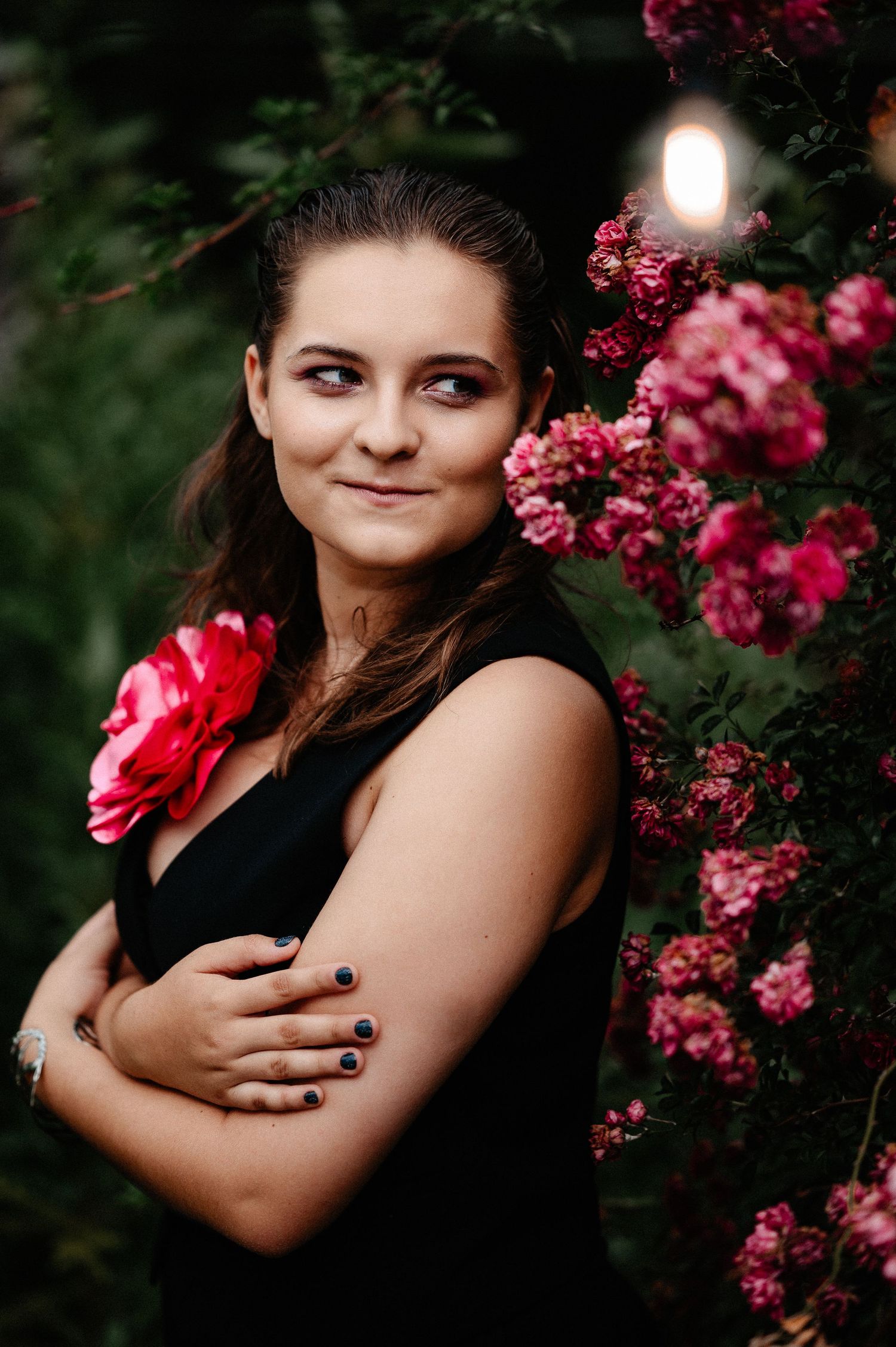 Smiling young woman in black sleeveless dress with pink rose corsage posing beside blooming rose bush.