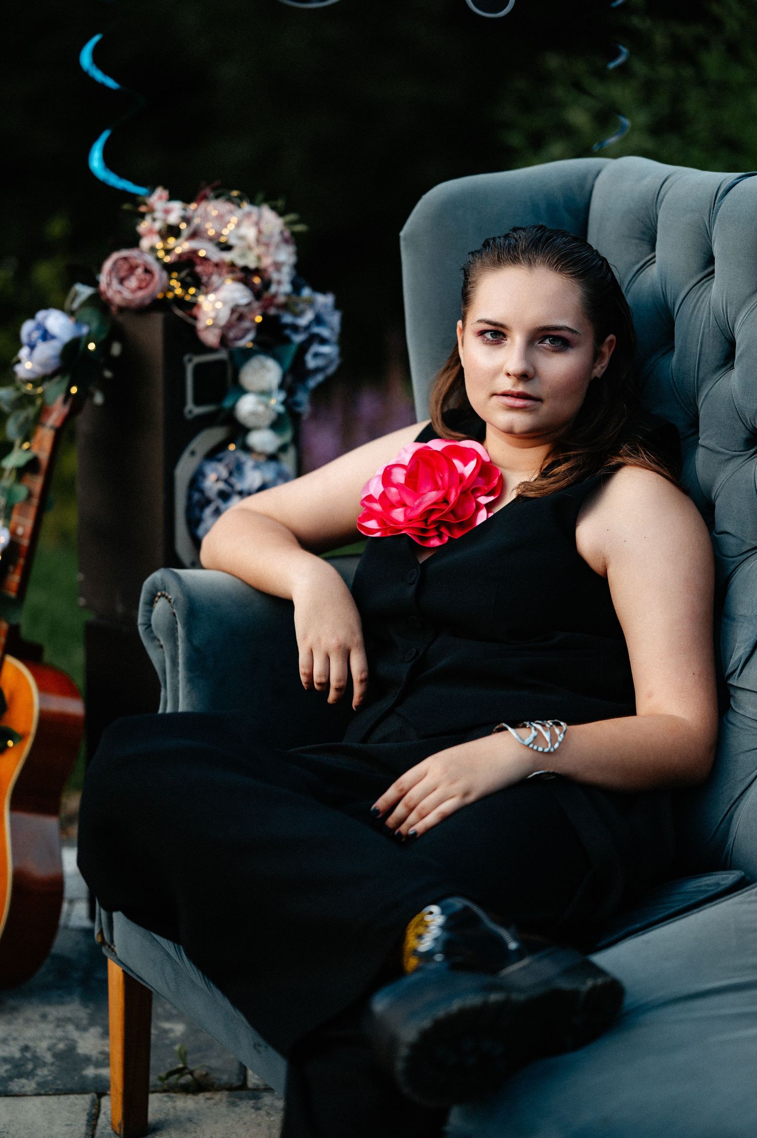 Young woman in black jumpsuit with pink flower corsage sitting confidently in a gray tufted armchair.