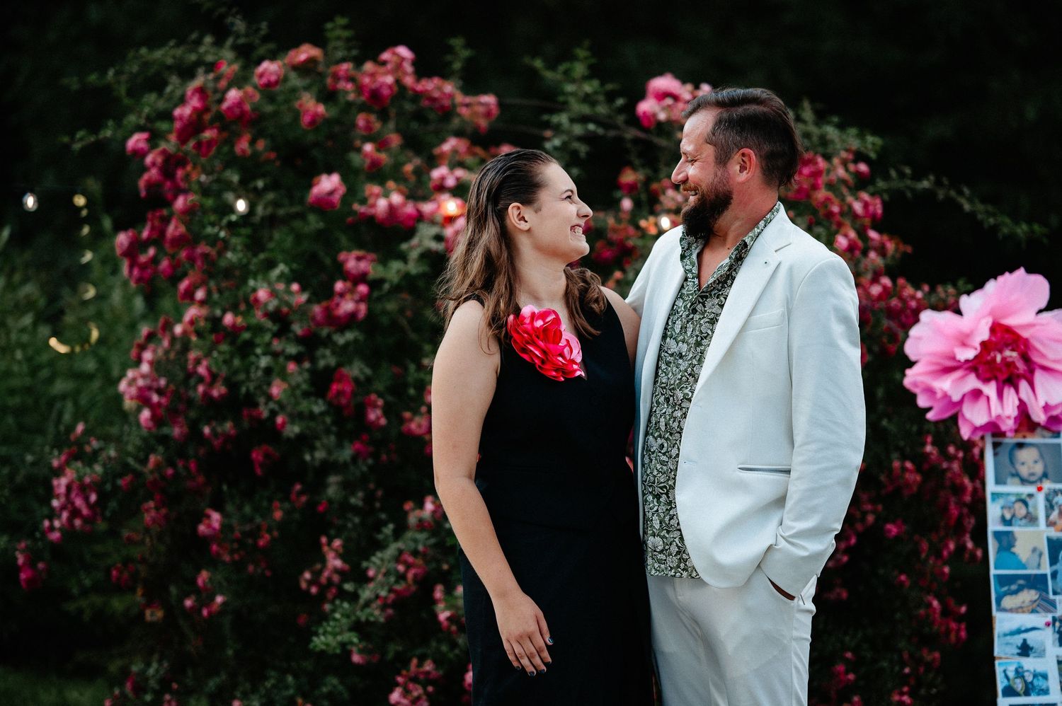 Man in white suit and woman in black dress sharing a laugh in front of pink rose bushes.