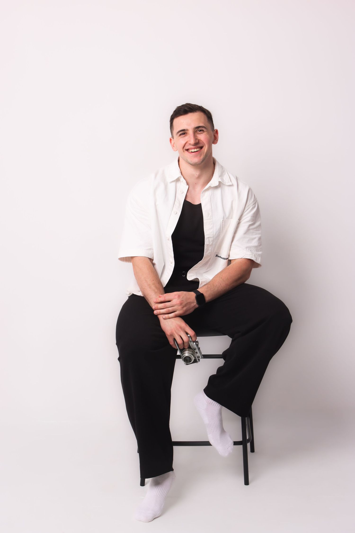 Smiling young man wearing white button shirt and black pants seated on stool with camera against white wall.