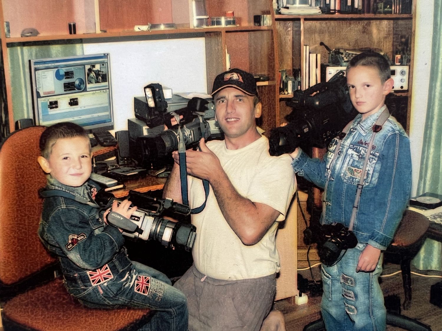 Man and two young boys posing together holding professional cameras and video equipment in home studio.