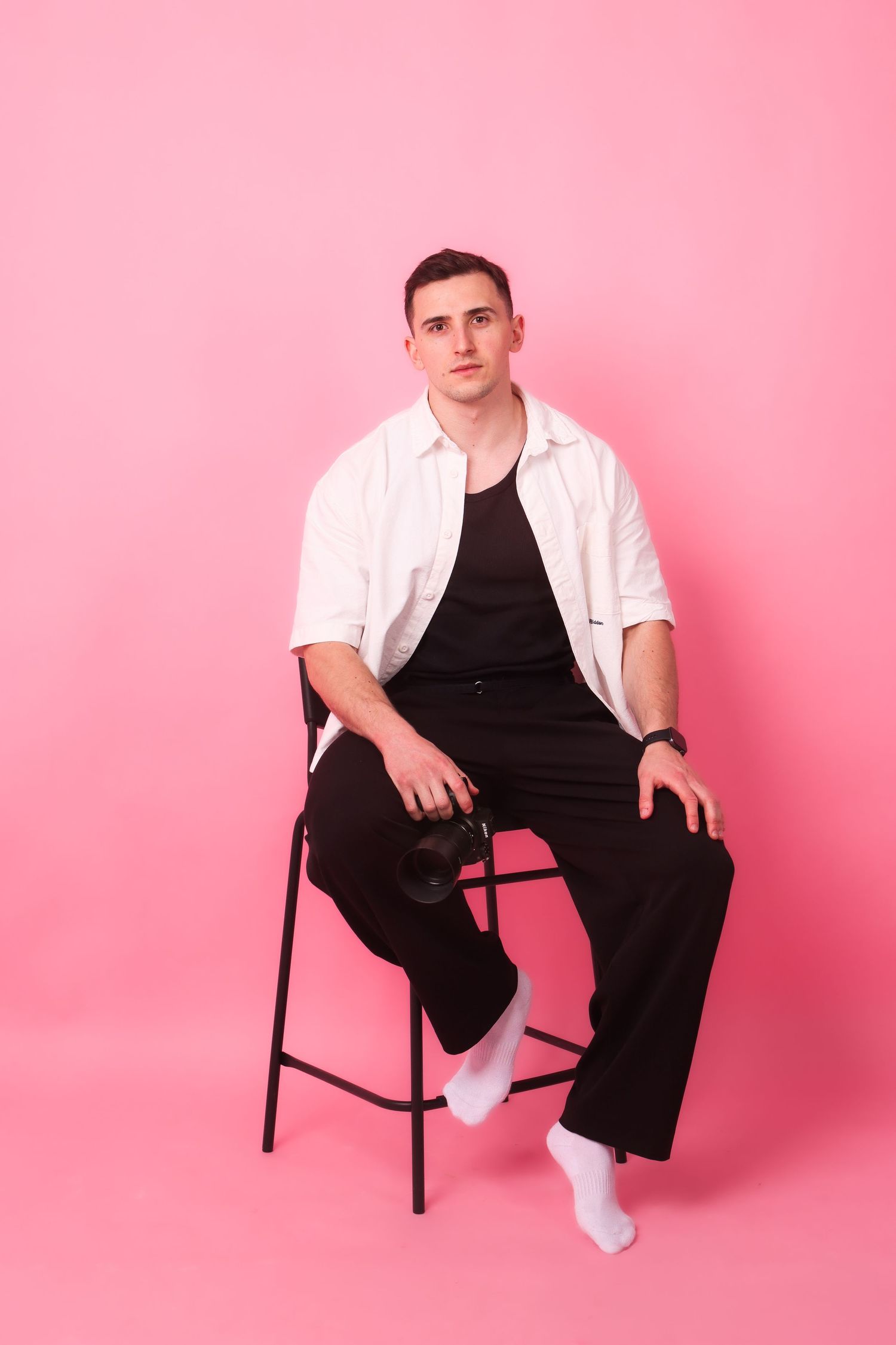 Young man in white shirt and black pants sitting on a chair against a pink background.