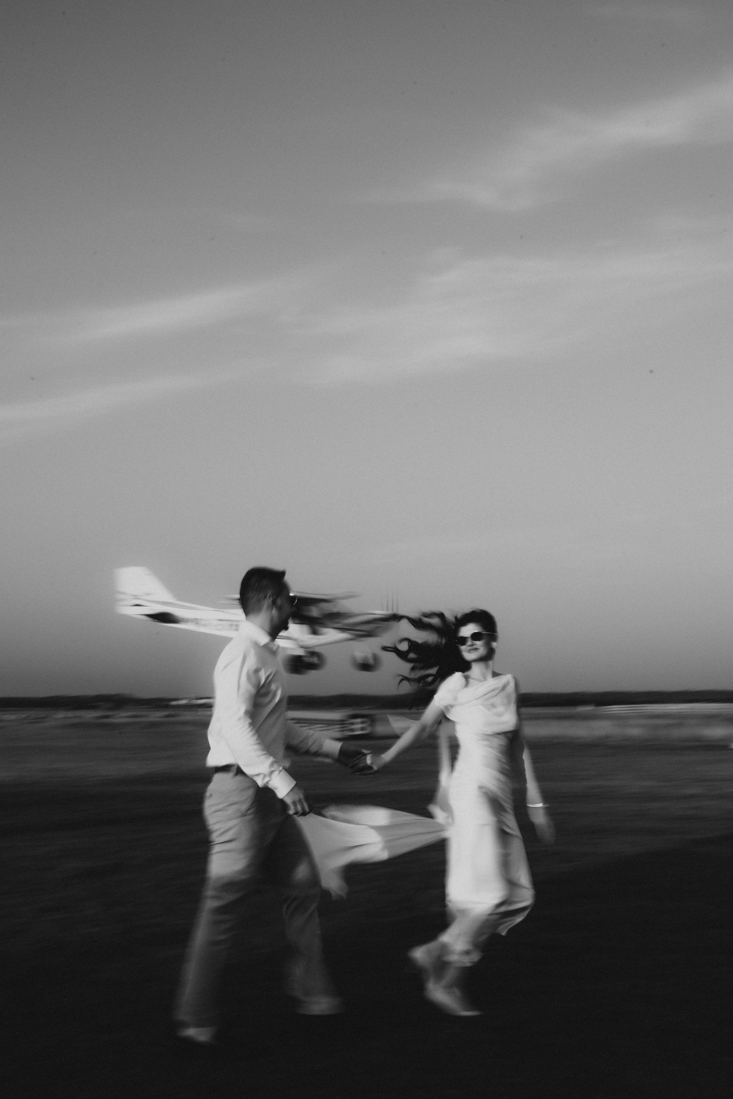 Black and white photo of a couple holding hands walking on a beach at sunset, woman in white dress with hair blowing.