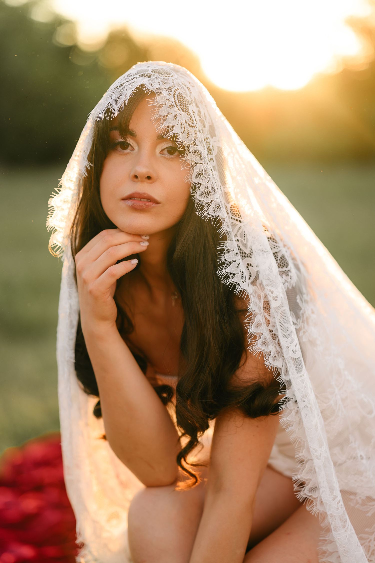A bride in a lace veil sits gracefully outdoors at golden hour, her dark hair flowing beautifully.