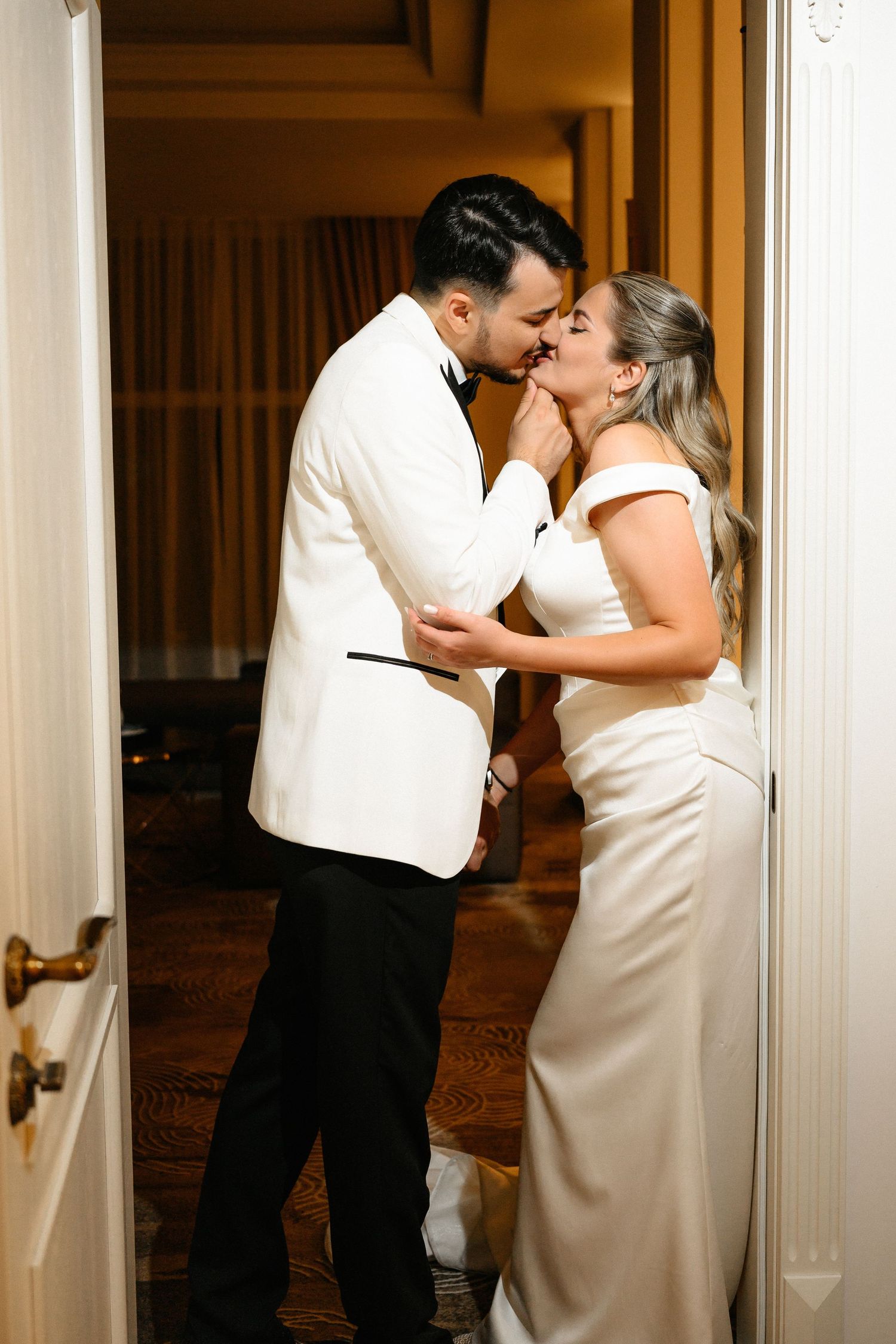 A bride and groom share a romantic kiss in a doorway, dressed in elegant white wedding attire.