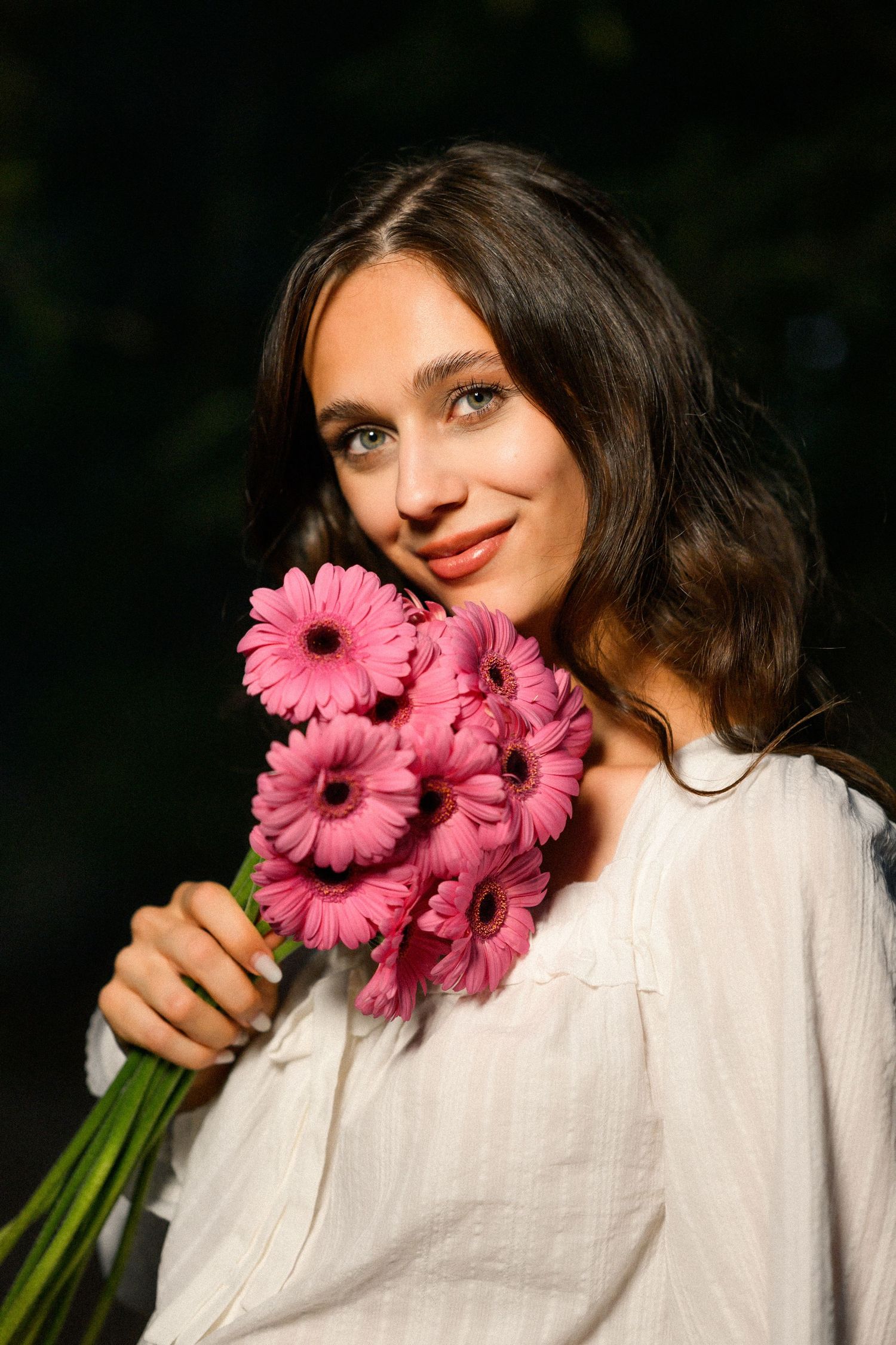 Young woman with wavy brown hair holding pink gerbera daisy bouquet, wearing white blouse outdoors.