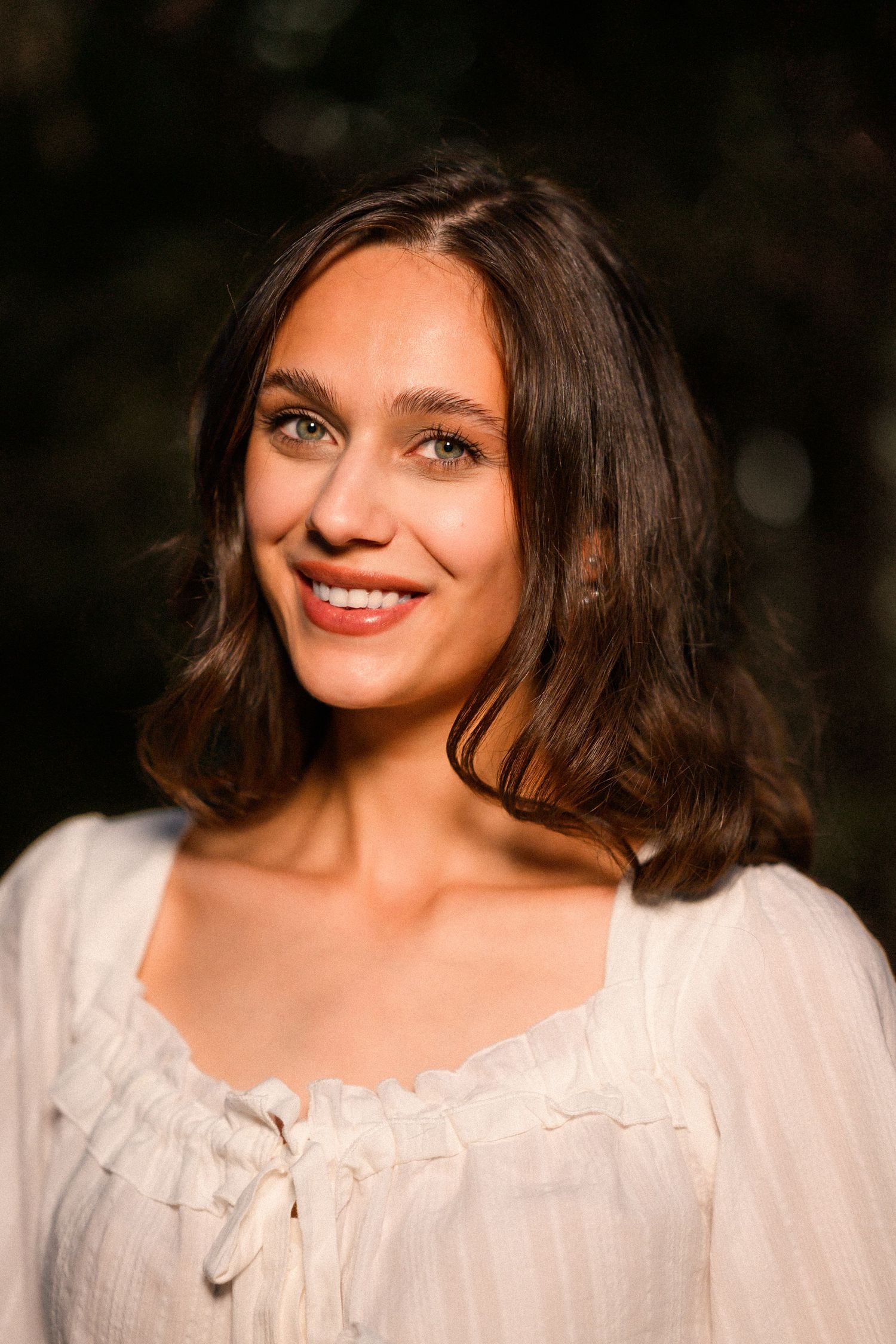 Young woman with dark wavy hair smiling outdoors, wearing a white lace blouse in warm golden sunlight.