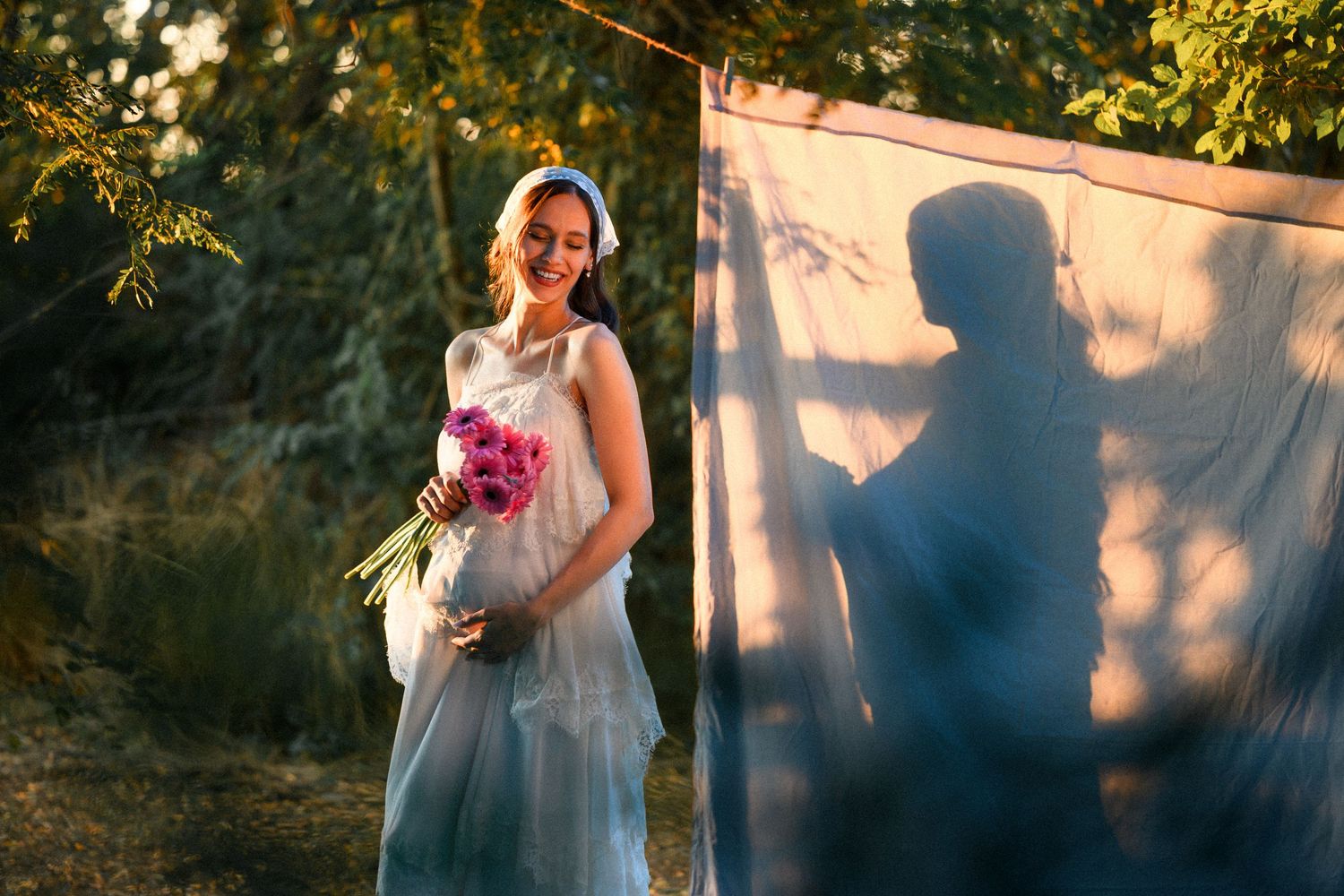 Smiling pregnant woman in white dress holding pink flowers outdoors, with her shadow cast on a white sheet backdrop.