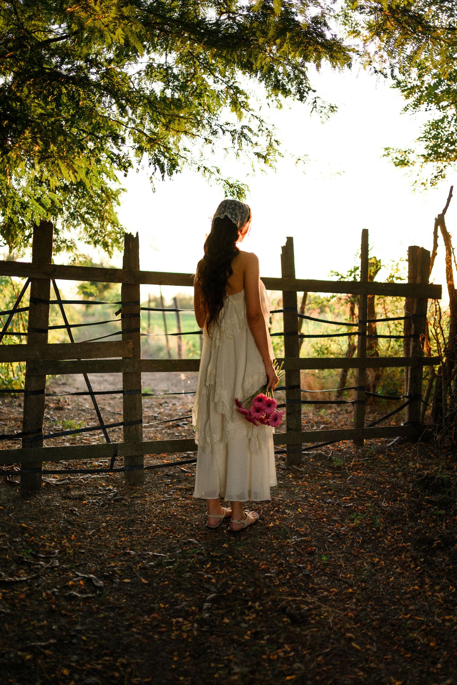 Woman in white dress holding pink flowers stands before rustic wooden fence in golden hour sunlight.
