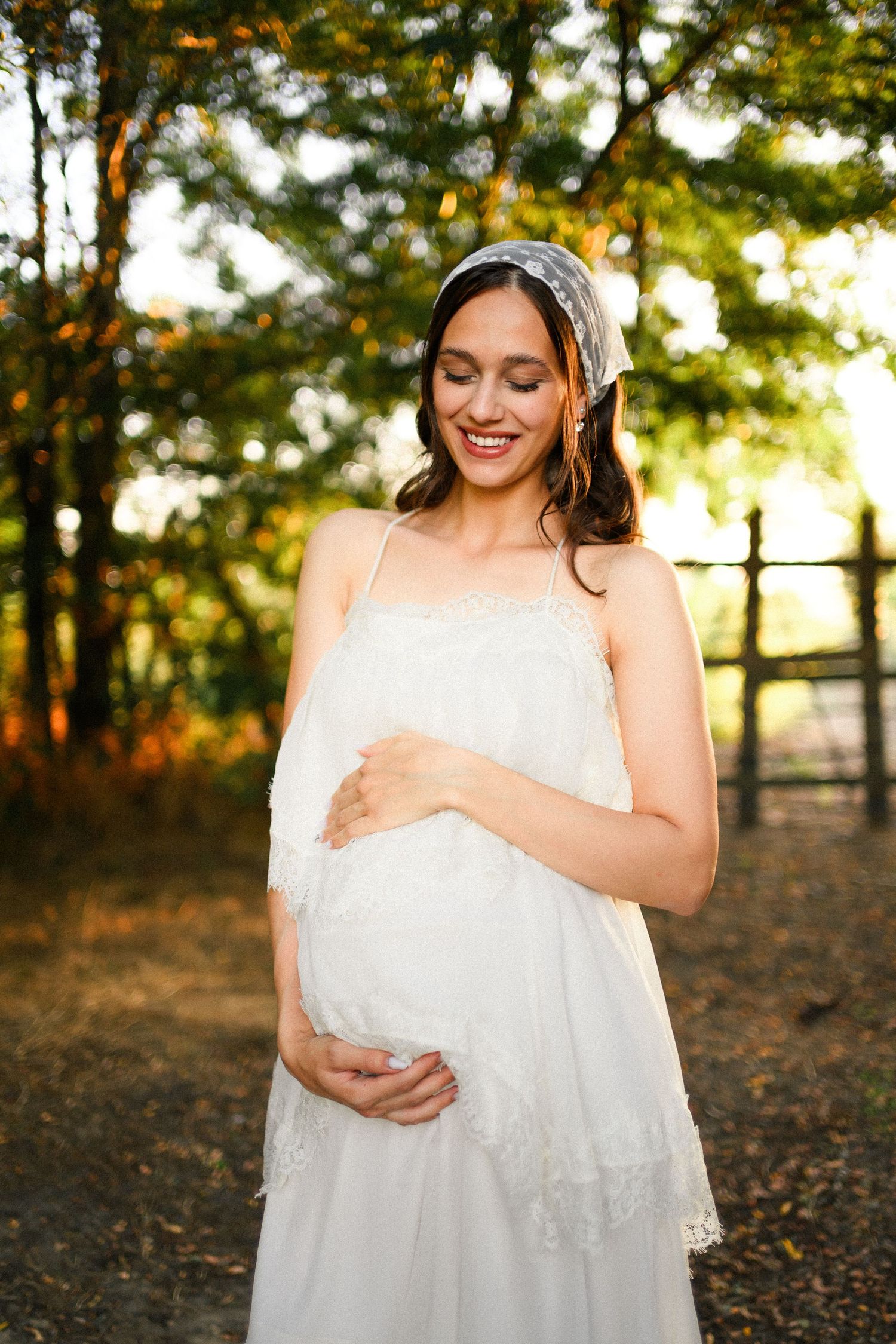 Smiling pregnant woman in white lace dress and veil headband cradling baby bump outdoors in golden sunlight.