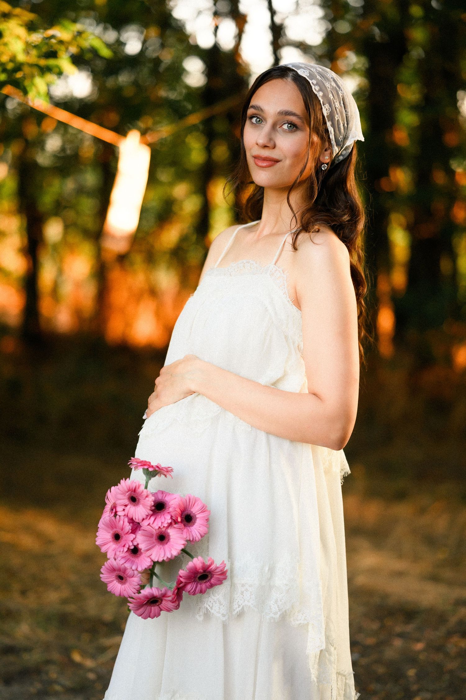 Pregnant woman in white dress holding pink gerbera flowers, smiling outdoors in golden sunset light with forest background.
